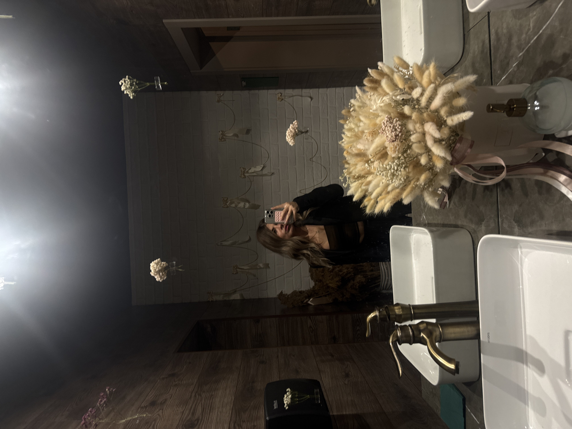 A person is taking a selfie in a bathroom mirror. The bathroom has wood-paneled walls and a white brick wall with decorative wall art. Two white sinks are visible, each with a brass faucet. A bouquet of dried flowers is placed on the counter. The city of Chaikovsky, Russia is not discernable from the image.