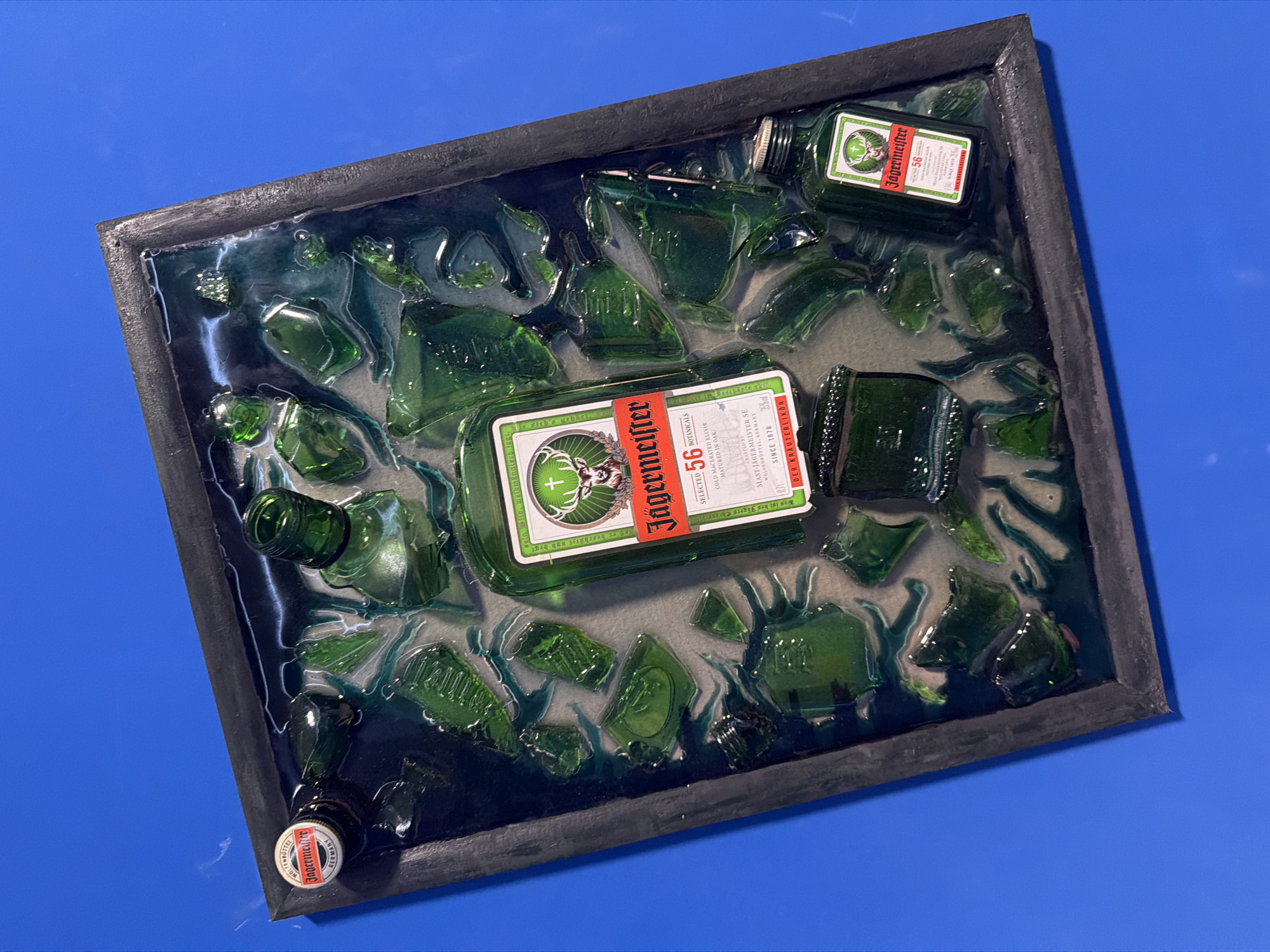 This image presents a framed mixed-media art piece, primarily featuring green glass Jägermeister bottles and their broken shards, set within a translucent, resin-like substance. The rectangular frame is dark, appearing black or very dark gray, with a slightly rough texture, and is displayed on a uniform, bright blue background.
The central elements within the frame are two Jägermeister bottles. A larger, mostly intact bottle rests prominently in the center, displaying its full label. A smaller, miniature Jägermeister bottle is situated in the upper right quadrant. Numerous fragments of broken green glass, consistent with Jägermeister bottles, are scattered throughout and embedded in the clear and semi-translucent resin. The resin itself exhibits variations in color, with darker, swirling patterns, possibly in deep green or teal hues, alongside clearer sections. A white and red Jägermeister bottle cap is visible in the bottom left corner of the frame.
No activity is taking place; the image captures a static art object. The lighting is bright and even, highlighting the reflective surfaces of the glass and the glossy texture of the resin.
Visible text on the main bottle's label includes "Jägermeister," the number "56," "SELECTED 56 BOTANICALS," "COLD MACERATED ELIXIR," "MATURED IN OAK," "CRAFTED BY MAST-JÄGERMEISTER SE WOLFENBÜTTEL, GERMANY SINCE 1878," and "DER KRÄUTERLIKÖR." A partial phrase "Was ist des Jägers Ehrens" is also visible. The smaller bottle's label also clearly shows "Jägermeister" and "56." The bottle cap features the Jägermeister logo and "GERMANY."