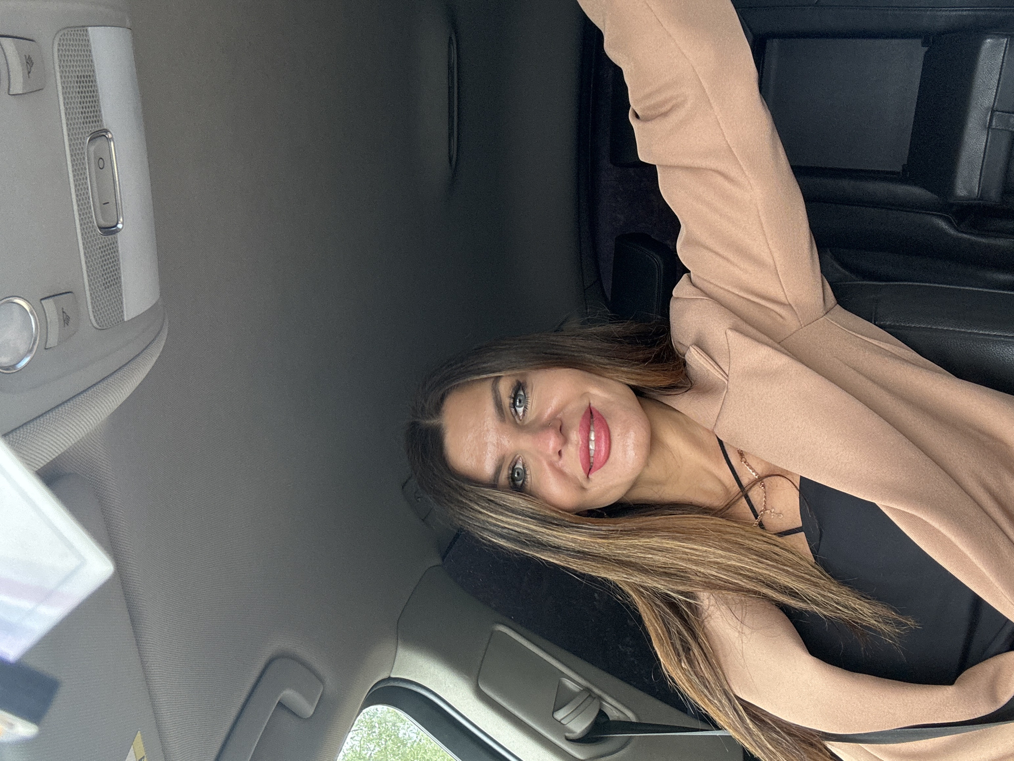 A young woman is depicted seated inside a car, looking directly at the camera with a pleasant, subtle smile. Her long, light brown hair with subtle highlights falls over her shoulder. She has striking blue eyes, defined eyebrows, and is wearing bright pink/coral lipstick, along with a small nose piercing. She is dressed in a black top layered with an open, light brown or beige blazer, and a delicate gold-colored necklace is visible around her neck.
The setting is a car interior, characterized by a gray, textured headliner and black car seats. An overhead console is prominent in the upper left, featuring a speaker grille and several buttons. One button clearly displays the numerals '0' and '-', likely for light or volume control. Natural daylight illuminates the scene, suggesting it is daytime. The image appears to be a casual portrait or selfie taken within a modern vehicle. This media file was captured in Chaikovsky, Russia.
