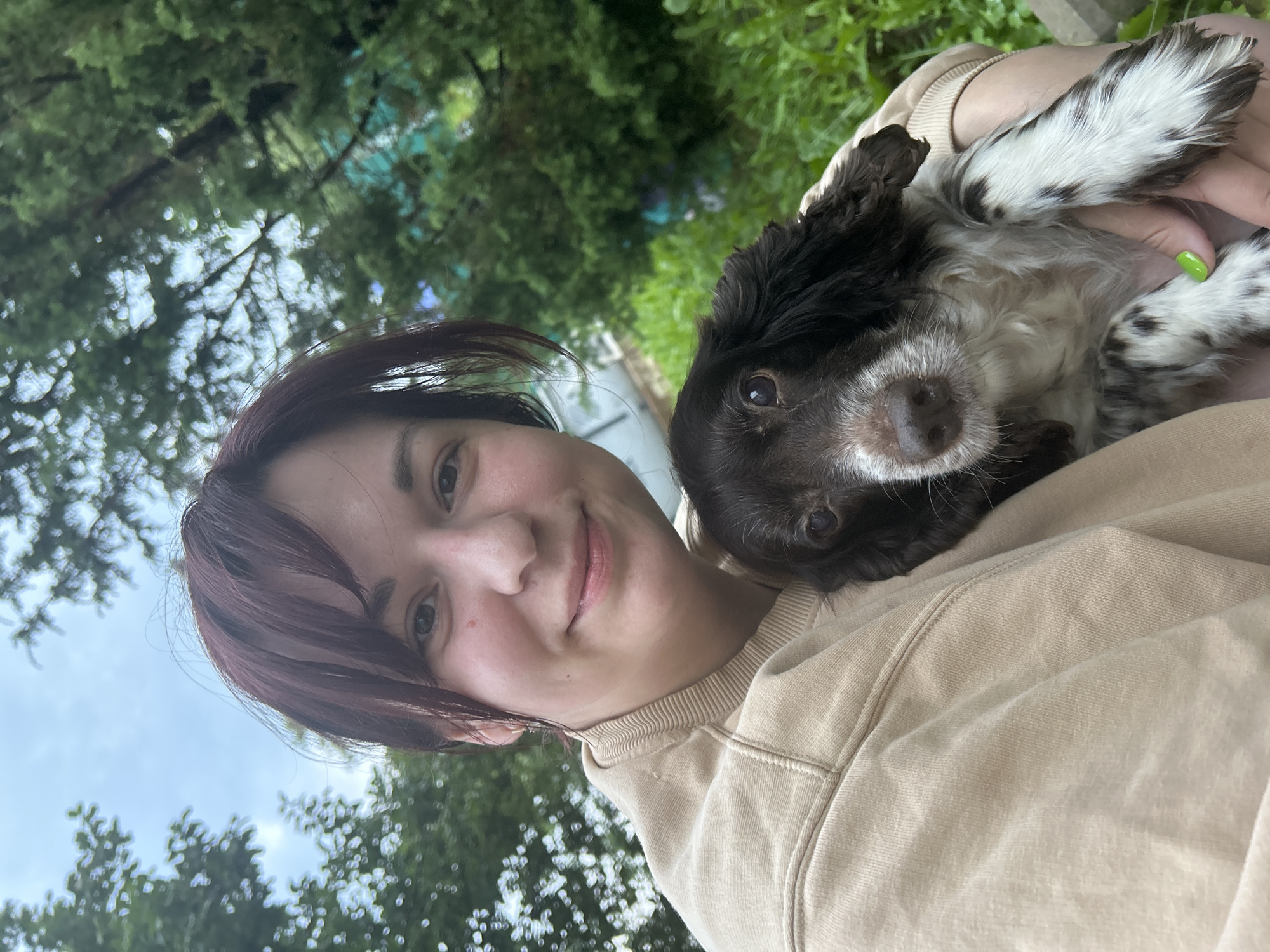 A young woman with short, dark red hair and a light complexion smiles as she holds a brown and white Springer Spaniel in her arms. She is wearing a beige sweatshirt. The dog is looking at the camera with its head resting on the woman's shoulder. They are outdoors with lush green foliage and trees in the background, suggesting a park or garden setting. The sky is overcast. The location in the city of Фоки, Russia, is not discernible from the image.