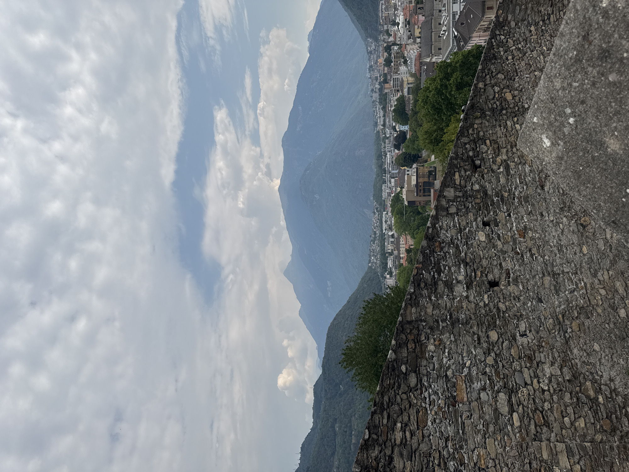 This image depicts a view from a high vantage point, likely within the historic Castel Grande in Bellinzona, Switzerland. The foreground is dominated by a textured, ancient stone wall constructed from irregular grey and brown stones of various sizes, suggesting a fortified structure. A lighter grey, weathered concrete object is visible in the lower right corner, also part of the vantage point.
Beyond the wall, a city unfolds with a dense arrangement of buildings featuring terracotta roofs and a mix of modern and older architectural styles. Green trees are interspersed throughout the urban landscape.
In the background, a dramatic mountain range rises, its slopes covered in lush green vegetation that transitions to a hazy, muted blue as the mountains recede into the distance. The sky above is filled with a mix of white and grey clouds, with patches of light blue sky visible. The overall scene is one of historical architecture overlooking a modern city nestled within a majestic natural landscape. There are no people or animals visible in the image.
