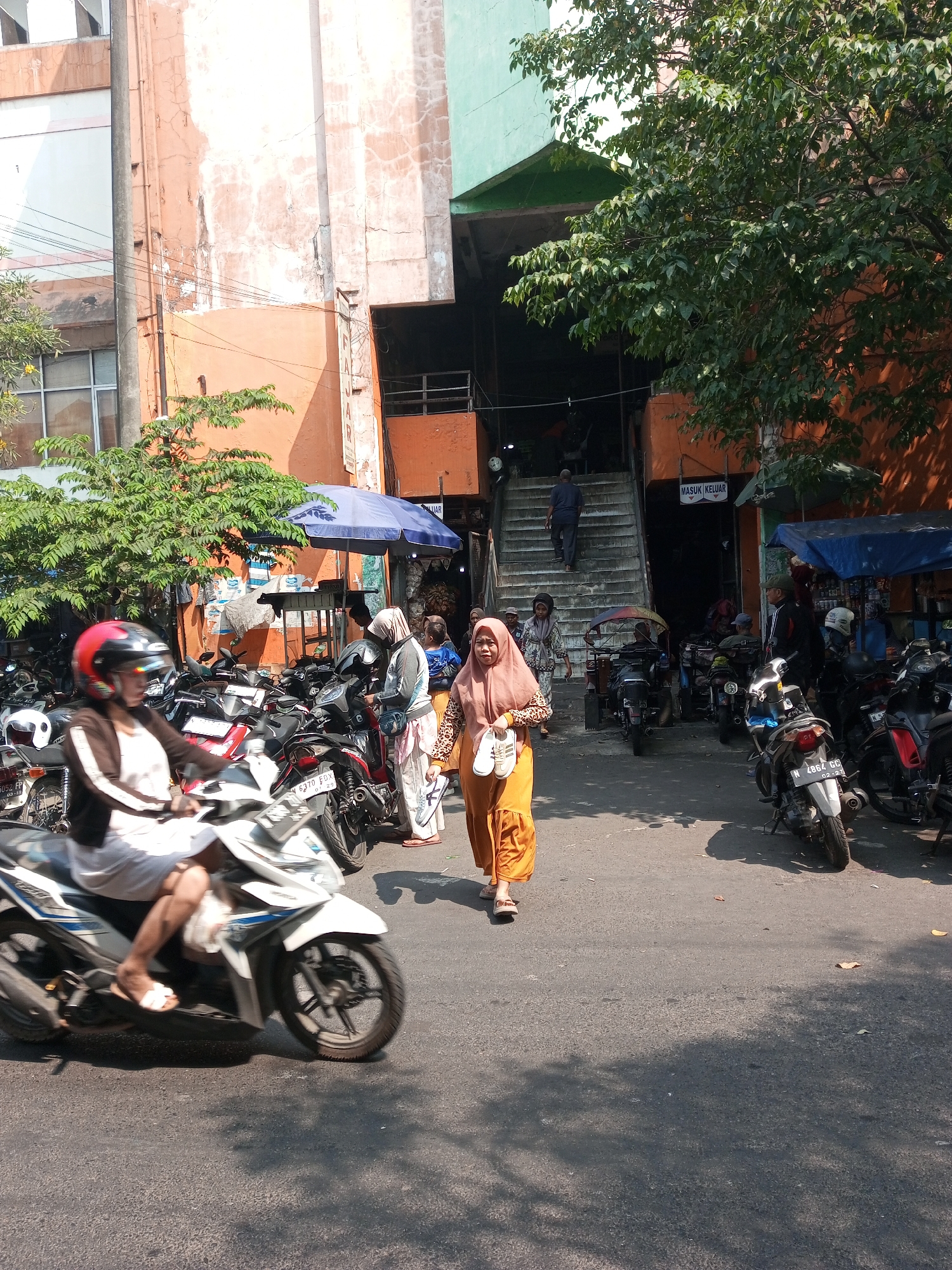 This image captures a bustling street scene in Malang, Indonesia, likely near a market or commercial area. The foreground is dominated by a person riding a white scooter, their legs bare and wearing white sandals. The scooter has blue and white detailing and a red and black helmet is visible on the rider's head. Behind the scooter, a row of parked motorcycles, predominantly black, fills the street.
In the mid-ground, several individuals are walking. A woman in an orange hijab and matching orange skirt walks towards the right, carrying a white bag and a pair of white shoes in her hand. Next to her, another woman in a light-colored hijab and patterned skirt also walks. To their left, a person in a grey hijab and patterned skirt stands near some parked motorcycles. Further back, a man in a blue shirt ascends a wide concrete staircase.
The environment features the weathered facade of an orange building on the left, with visible cracks and peeling paint. Adjacent to it is a greenish building. A large tree with lush green foliage casts shadows across the asphalt. Blue and patterned awnings provide shade over stalls or shops. The background reveals more structures and parked vehicles. Signs are visible, including one with "MASUK KELUAR" (Enter Exit) in English, indicating a public space. The overall impression is a vibrant, active urban environment under bright sunlight.