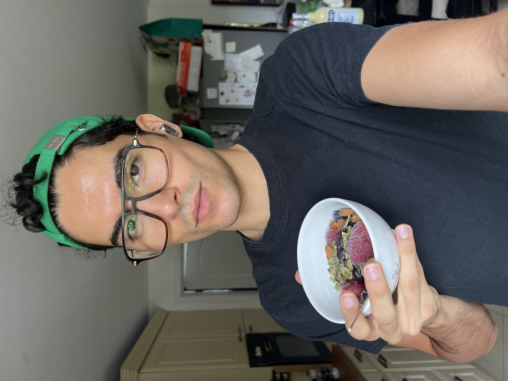 A person wearing a black t-shirt, glasses, and a green baseball cap is holding a white bowl filled with berries, seeds, and almonds. The person is looking at the camera. The background shows a kitchen with cabinets, a refrigerator, and bottles. There is no indication of the location being Rotheux-Rimière, Belgium.