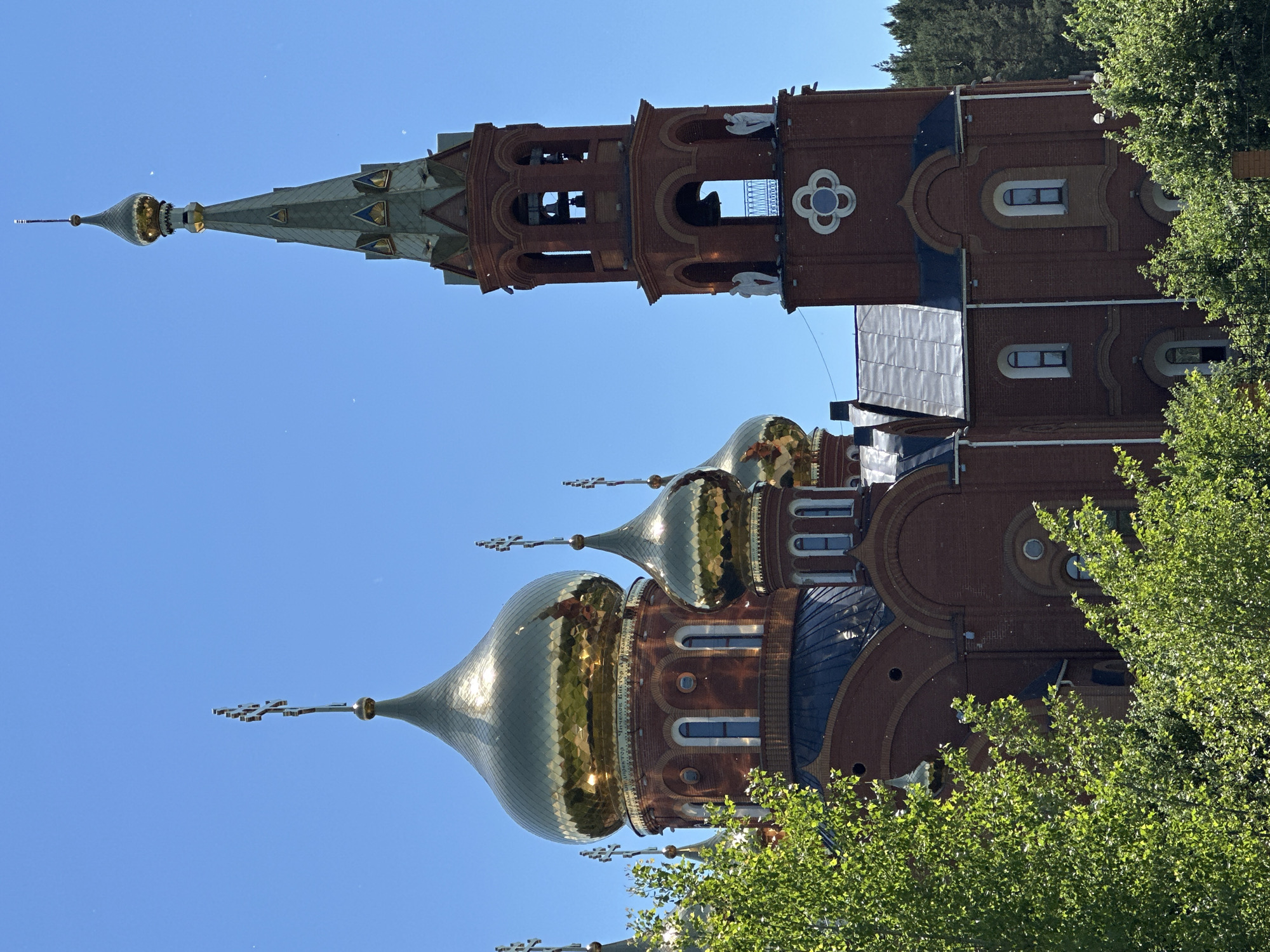 The image shows a large, red brick Orthodox church with several golden domes and a tall bell tower. The church has arched windows and intricate architectural details. Green trees and foliage are visible in the foreground, partially obscuring the lower portion of the building. The sky is a clear, bright blue. The bell tower features arched openings where bells are visible and decorative statues of angels. The golden domes are topped with crosses. This is the Transfiguration Cathedral, located in Chaikovsky, Russia.