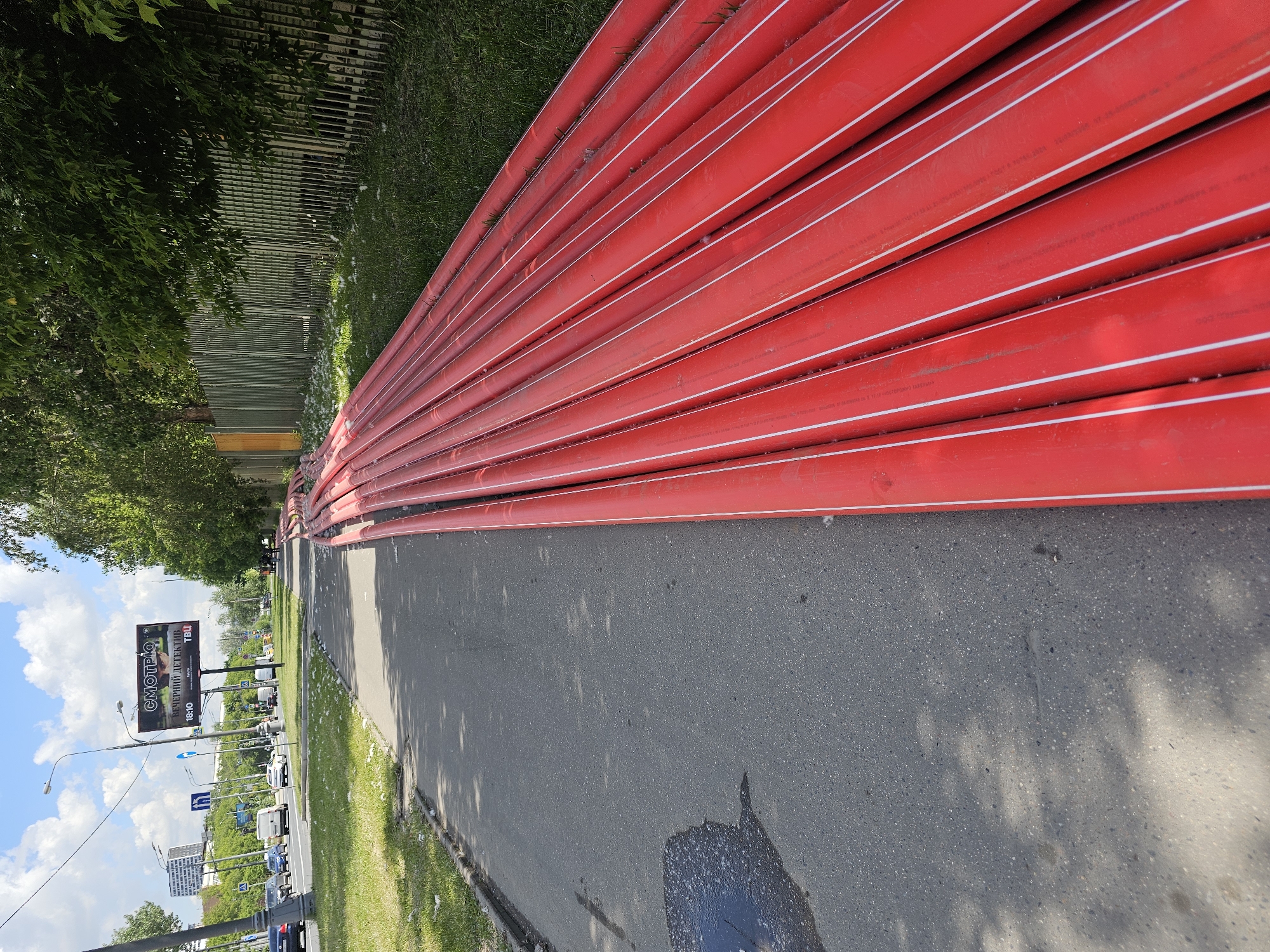 The image, oriented correctly, depicts a long stack of bright red pipes with thin white longitudinal stripes, lying horizontally on a grassy verge beside an asphalt road. These pipes, likely made of HDPE, bear faint white text including "SDR 17", "PN10", and "PE100", indicating their material and pressure rating.
To the left of the pipes, a narrow strip of green grass borders the asphalt road. The road surface shows variations in shade and texture, with shadows from overhead trees dappling its surface. Further along the road, a dark, wet patch is visible.
On the opposite side of the road, a busy multi-lane street is visible with cars and other vehicles in motion. Streetlights and road signs are present, and a large billboard with a dark background stands prominently. The billboard features Cyrillic text: "СМОТРЮ" (SMOTRYU - I'm watching/looking) in large letters, and "ВЕЧЕРНИЙ ДЕТЕКТИВ" (VECHERNIY DETEKTIV - Evening Detective) below it, along with a "TBJ" logo and the number "1880".
Beyond the road and billboard, a dense line of green trees forms a natural barrier, and a grey metal fence can be seen through their foliage. In the far distance, urban buildings and structures are visible under a bright blue sky dotted with fluffy white clouds.
The scene suggests an ongoing infrastructure project, possibly related to water or gas supply, with pipes awaiting installation. It is daytime, characterized by bright sunlight and clear weather, consistent with the discernible shadows and bright sky. The Cyrillic text on the billboard confirms the location in Moscow, Russia. No people are visible, and no specific event is taking place beyond general urban activity and the presence of construction materials.