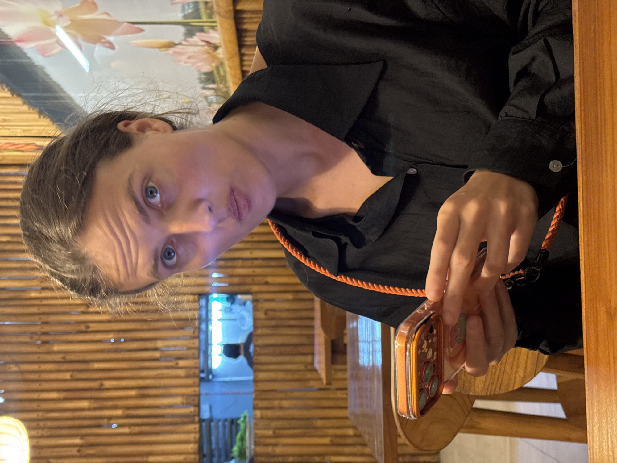 A woman is sitting at a wooden table, holding an orange phone with a matching orange cord. She is wearing a black shirt. The background consists of a bamboo wall and a glimpse of a building exterior with blue and white elements, possibly indicating the location of the image is in Kuta Selatan, Indonesia.