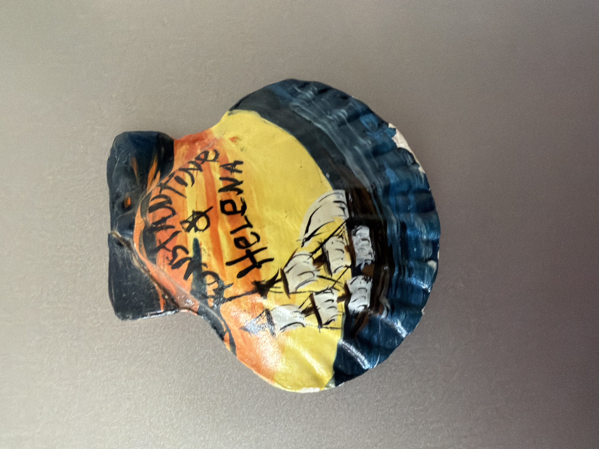 The image shows a scallop shell ornament painted with a sailing ship against a sunset sky and ocean. The text "CONSTITUTION & HELENA" is written in black on the shell. The shell is resting on a reflective, grey surface, likely a refrigerator door. The background is a plain, neutral color. There are no people or animals in the image. The ornament is likely a souvenir. There is no information in the image to suggest a location in Pazardzhik, Bulgaria.