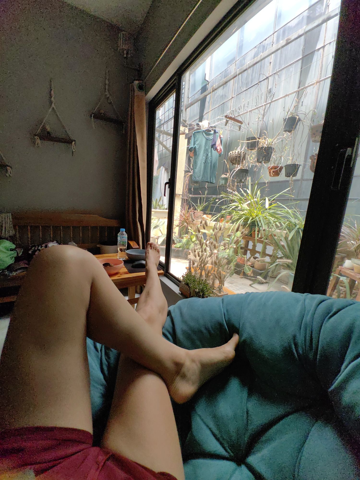 The image depicts a person relaxing indoors, seemingly in a residential setting in Hà Nội, Vietnam. The person is seated in a deep turquoise armchair or large cushion, with their legs crossed and extended towards the viewer. They are wearing what appears to be a maroon-colored garment, possibly shorts.
In the background, there is a room with a wooden couch, a small wooden table on which sits a water bottle, a pink bowl, and another bowl. The room is somewhat dimly lit, contrasting with the bright outdoor area visible through a large sliding glass door/window.
The exterior space beyond the glass features a vertical garden or a courtyard with numerous potted plants hanging from wires and arranged on shelves. Clothes hanging on a clothesline are also visible. The overall impression is of a relaxed, indoor/outdoor living space in an urban environment.
