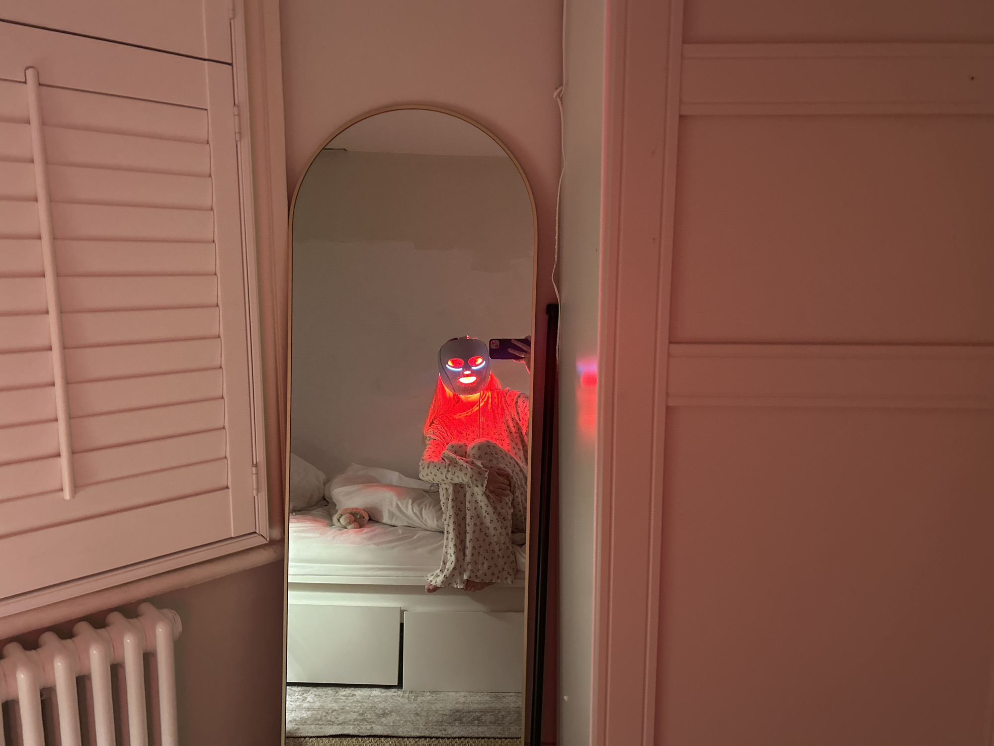 The image displays an interior residential scene, specifically a bedroom in London, United Kingdom, evidenced by a classic white column radiator and louvered window shutters. A full-length, arch-top mirror with a light-colored frame centrally reflects an individual. This person is seated on a white bed with knees drawn towards the chest, wearing light-colored floral pajamas. A grey light therapy mask, emitting intense red light, covers the person's face, illuminating the mask, the upper body, and casting a reddish glow on the immediate reflection. A dark-colored smartphone is held in the person's right hand, angled as if capturing a mirror selfie. On the bed, a small, light-colored plush toy is visible near a pillow. The white bed frame incorporates two storage drawers. White walls, a segment of a light-colored patterned rug, and a white electrical cable vertically mounted beside the mirror complete the visible elements of the environment.