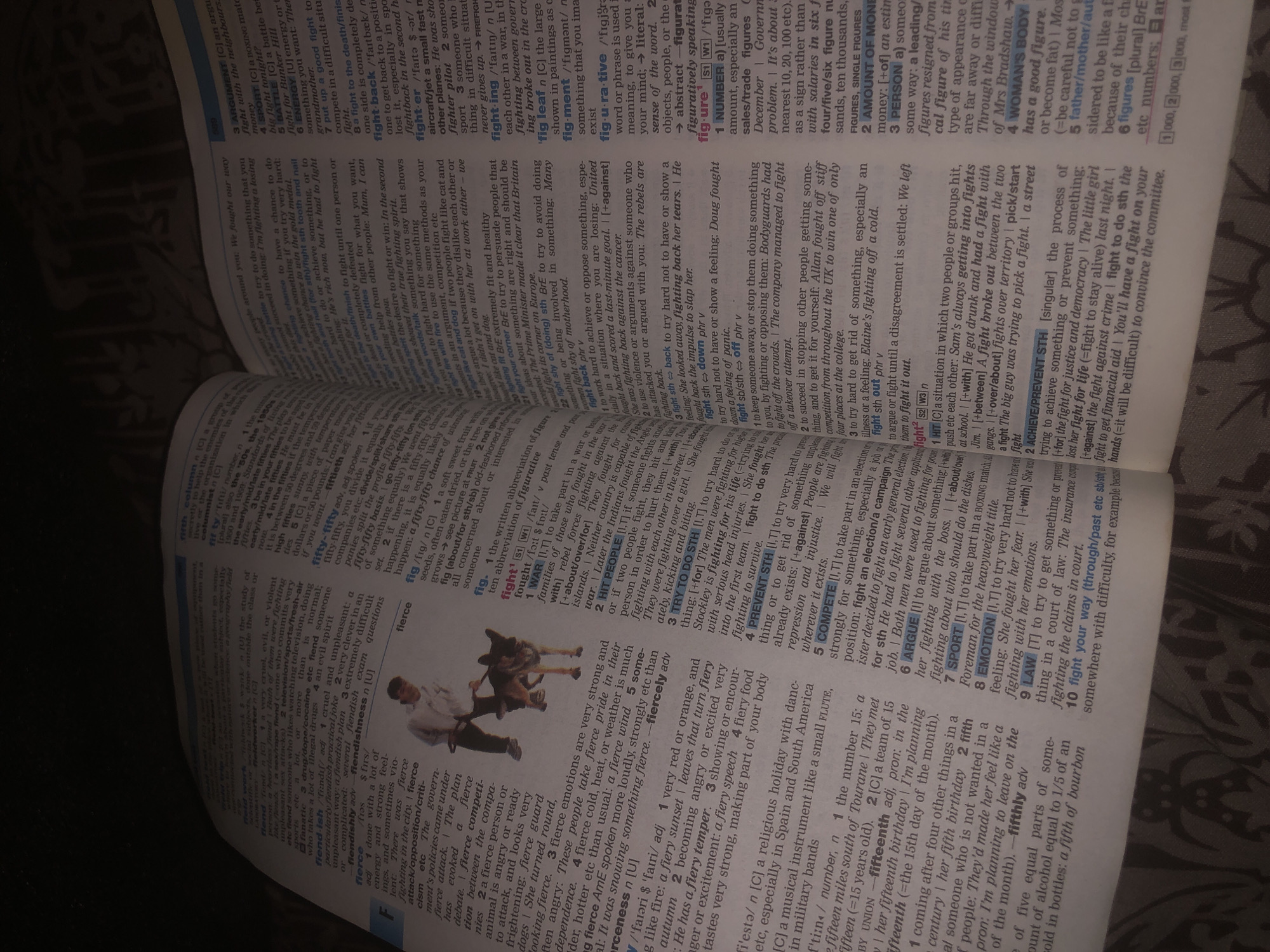 The image shows a close-up of an open dictionary. The primary subject is the text within the dictionary, with a blurry background suggesting it's a casual setting, possibly a home or library. The text is dense and organized into entries with definitions and examples.
Several entries are visible, including "fight", "figment", "fifty-fifty", "fifthly", and "fierce". The entries for "fight" offer multiple meanings and contexts, such as physical combat, arguments, and political struggles. The definition of "fierce" describes strong emotions and is illustrated with examples of passionate individuals. The word "fifty-fifty" is defined as an equal division of something, and "fifthly" is an ordinal number.
Notable details include the small print for citations and cross-references, the numbered sub-entries, and the occasional inclusion of phonetic transcriptions. The lighting suggests it might be daytime, but there are no strong indicators of time of day. The setting is neutral, and no specific weather is implied. The overall impression is one of information retrieval, focusing on the linguistic and etymological details of the English language.