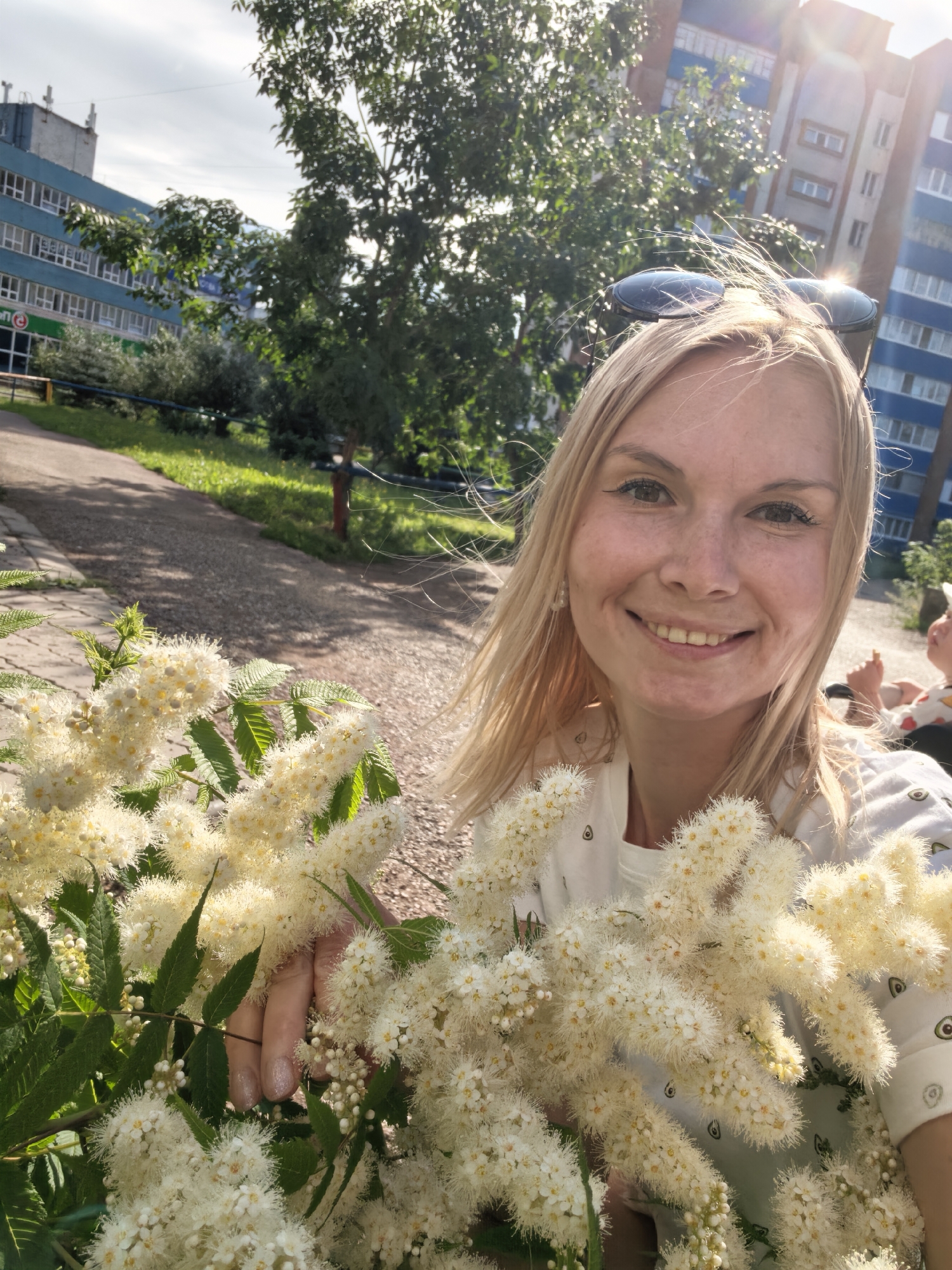 A blonde woman is taking a selfie outdoors in a city park in Sterlitamak, Russia. She is holding a large bouquet of fluffy white flowers. The woman is smiling at the camera, and her sunglasses are perched on her head. Behind her, there are apartment buildings, trees, and a paved walkway. The sun is shining, creating lens flare in the upper right corner of the image. In the background, a child can be seen sitting in a stroller, and another person is partially visible. The woman is wearing a white t-shirt with an avocado print.