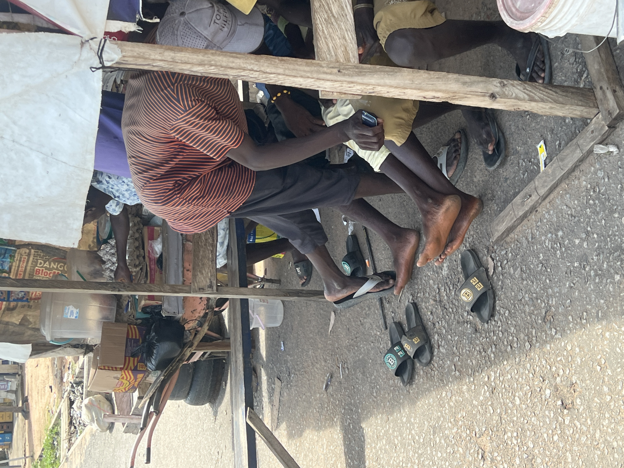 The scene depicts an outdoor roadside or market stall in Jalingo, Nigeria, under bright daylight. A makeshift wooden structure with a white fabric awning provides shade. Beneath the awning, a person wearing an orange and black horizontally striped short-sleeved shirt and dark shorts is bent forward. Several other individuals are partially visible, mostly showing their lower legs and feet, suggesting they are seated or standing under or near the stall. One person wears a grey baseball cap with "TOMMY HILFIGER" visible on the front. A dark-skinned hand holds a black flip phone, while another hand is near cardboard egg cartons.
On the wooden structure and ground, various items are present. Two large, clear plastic water dispensers, one with a blue label, are visible. Several cardboard boxes are stacked, one prominently displaying "DANGOTE BLOCK" branding. Cardboard egg cartons are arranged to the right. A black plastic bag rests on a wooden frame. On the ground, which appears to be worn asphalt or concrete, are three pairs of black slide sandals, two featuring a gold "BB" logo and one with a green "B" logo. Other footwear includes black flip-flops with white straps and black flip-flops. Scattered debris, including leaves and small pieces of paper, litters the ground. In the background, a two-wheeled cart frame with old tires leans against the stall, and beyond that, an unpaved reddish-brown dirt area with sparse vegetation borders a paved road. A white plastic bucket with a pink lid is in the lower right foreground.