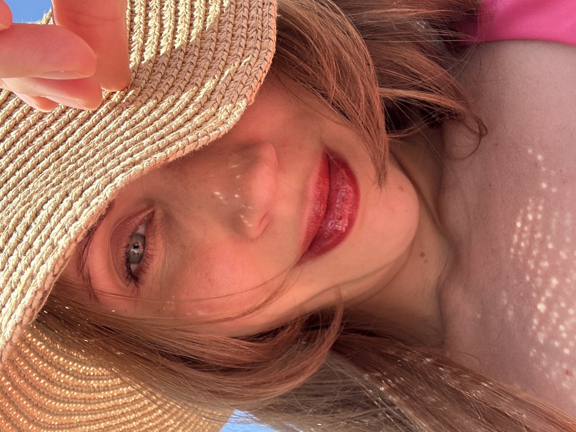 A close-up, tilted, low-angle shot captures a person wearing a straw hat. Part of the hat is held by a hand. The person has fair skin, light brown hair, and wears red lipstick. Sunlight filters through the hat, casting dappled light on their face. The background is a blurry blue, suggesting sky. There is no information suggesting Fethiye, Turkey.