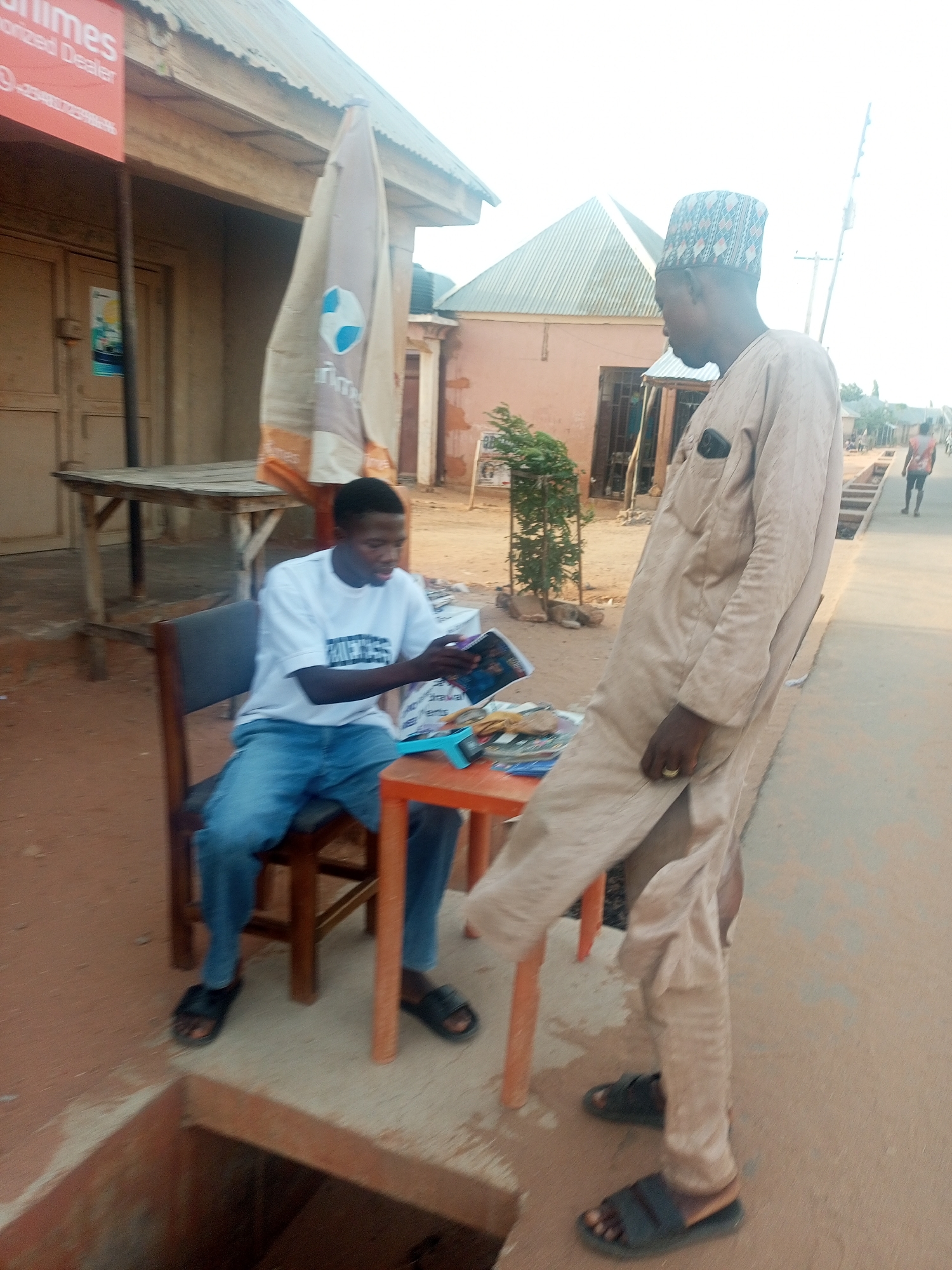 The image depicts an outdoor scene in Tempure, Nigeria. A young man sits on a chair at a small table, looking at a book or magazine. He is wearing a white t-shirt, blue jeans, and sandals. Standing next to him on the sidewalk is a man wearing a light-colored traditional outfit and a patterned cap. There is an umbrella with a logo behind the seated man. In the background are buildings with metal roofs and a person walking down the street. There is an open drain hole in the foreground.
