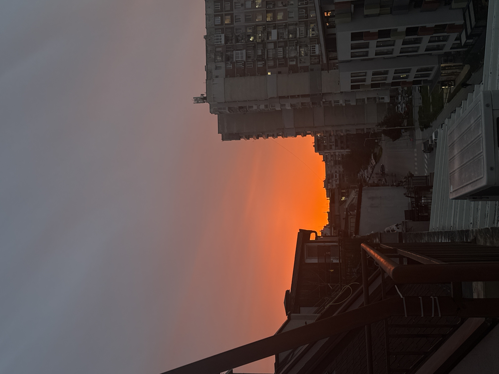 The image depicts an urban scene at dusk, likely in Varna, Bulgaria, characterized by densely packed buildings and a fiery orange sunset. A large apartment complex with numerous windows occupies the upper-right quadrant, its facade facing the viewer. Below, a street scene unfolds with buildings of varying heights and architectural styles. Hints of vehicular traffic are visible. The foreground features rooftops and metallic structures, possibly a fire escape or ventilation equipment. The sky transitions from a dark grey to a vibrant orange near the horizon. The overall tone is one of urban density bathed in the dramatic light of the setting sun. The image is rotated 90 degrees clockwise.