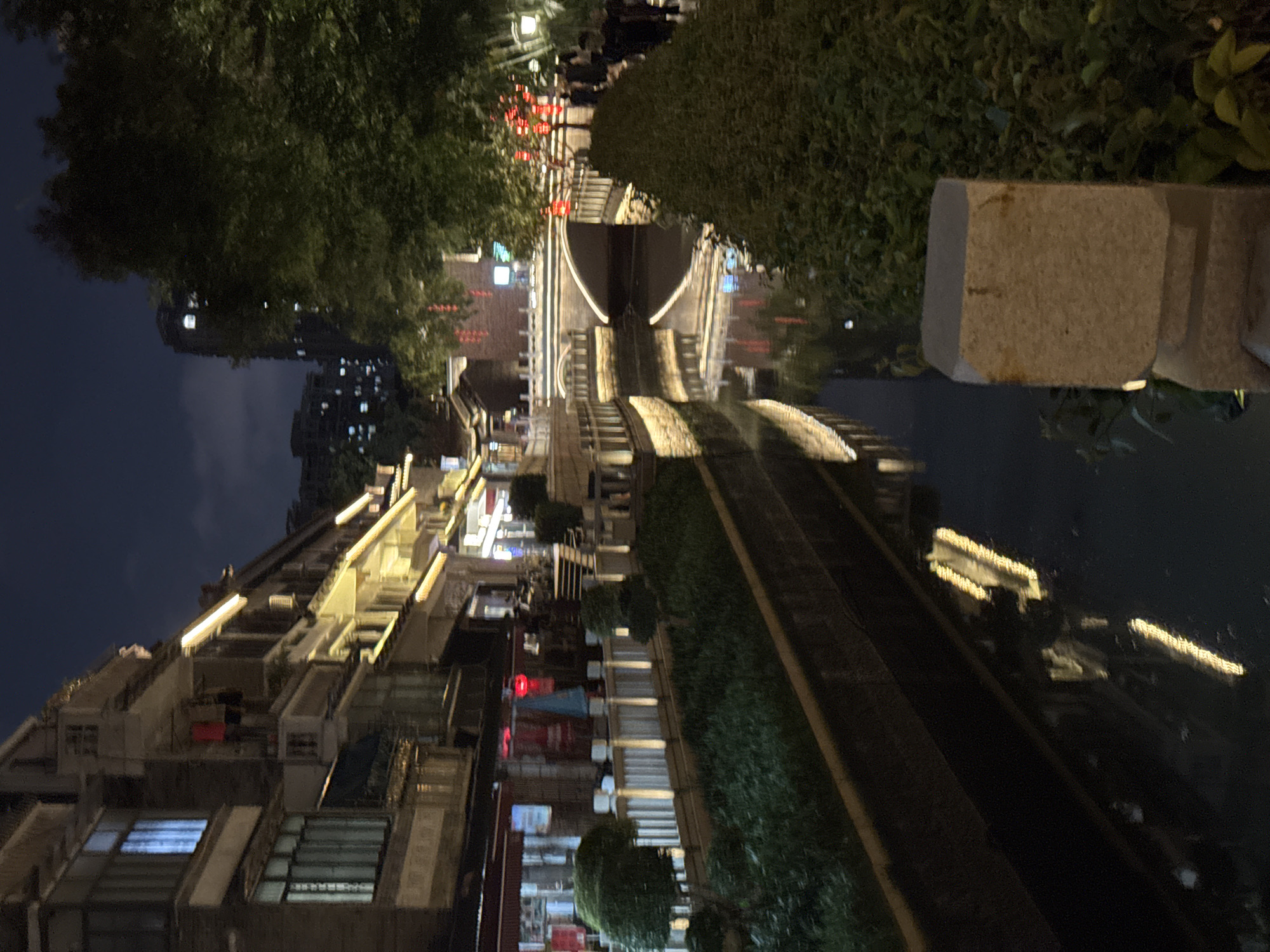 The photograph captures a night scene in Liwan District, China. It features a canal reflecting illuminated buildings, a bridge, and surrounding vegetation. The buildings are multi-storied and adorned with lights. The canal runs linearly through the scene with lights reflecting on its surface. A stone structure is visible in the foreground, while trees and dense shrubbery border the waterway. A bridge arches over the canal, and people are visible crossing at the top. The sky is a dark blue, indicating nighttime.