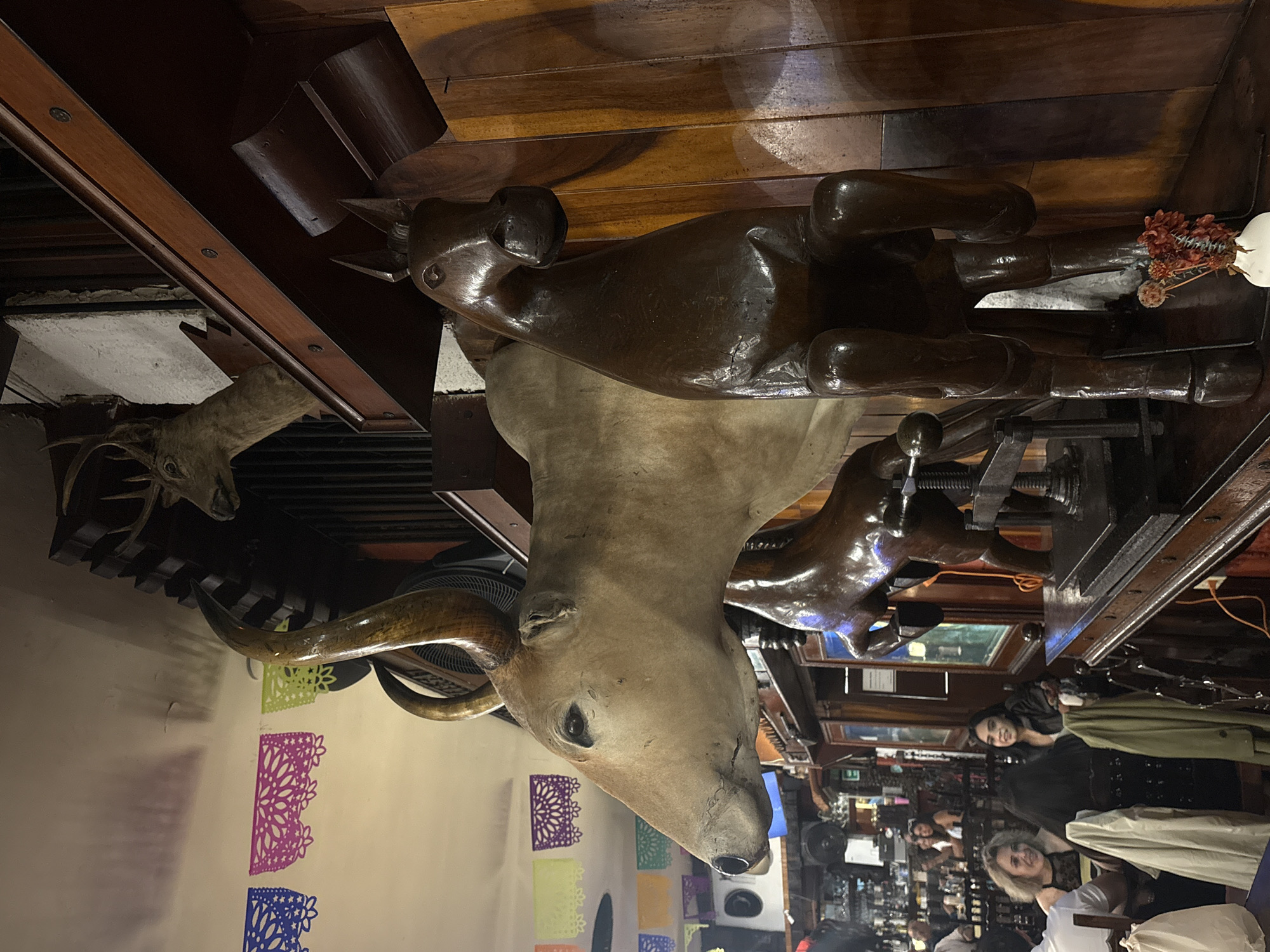 This image appears to be taken inside a restaurant or bar in Mexico City, Mexico. The foreground features taxidermied animal heads, including a bull and a deer, mounted on a wall. The wall is decorated with colorful, patterned paper cutouts. In the background, there are people, some of whom are seated at tables. Shelves stocked with bottles and other items are also visible in the background, suggesting a bar area. The restaurant seems to have a rustic or traditional decor, with wooden furniture and fixtures.