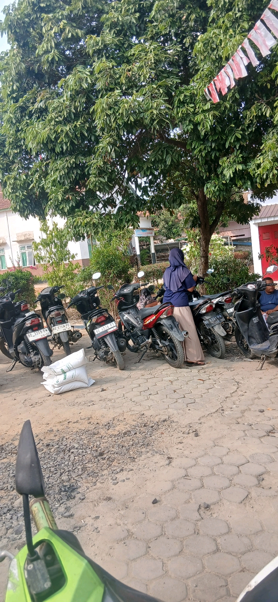 In Metro, Indonesia, a group of motorbikes are parked in a lot. A woman wearing a hijab and a long skirt stands by one of the motorbikes. In the background, a building with white walls and windows is visible. A large tree with green leaves provides shade over the scene. In the foreground, a green motorbike mirror is partially visible.