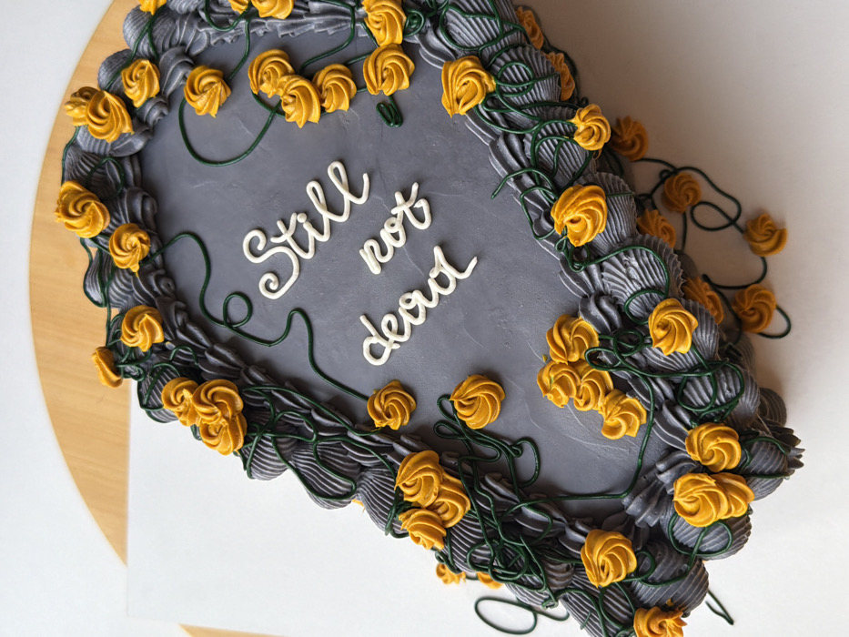 A rectangular cake, designed to resemble a tombstone, is depicted from a slightly elevated, angled overhead perspective. The cake features a smooth, medium-gray frosting on its top surface, bordered by an intricate, textured dark gray piped frosting pattern, suggesting carved stone or elaborate molding. Numerous small, vibrant orange-yellow frosting rosettes are meticulously arranged along this border and scattered across the gray surface. These rosettes are interconnected by thin, dark green piped lines, styled as winding vines or stems. Centered on the gray surface, white cursive lettering spells out "Still not dead." The cake is positioned on a flat, white surface, which itself rests upon a light brown wooden surface. The scene is static, devoid of people or direct actions, and is located in an indoor environment within the city of Tel-Aviv, Israel.