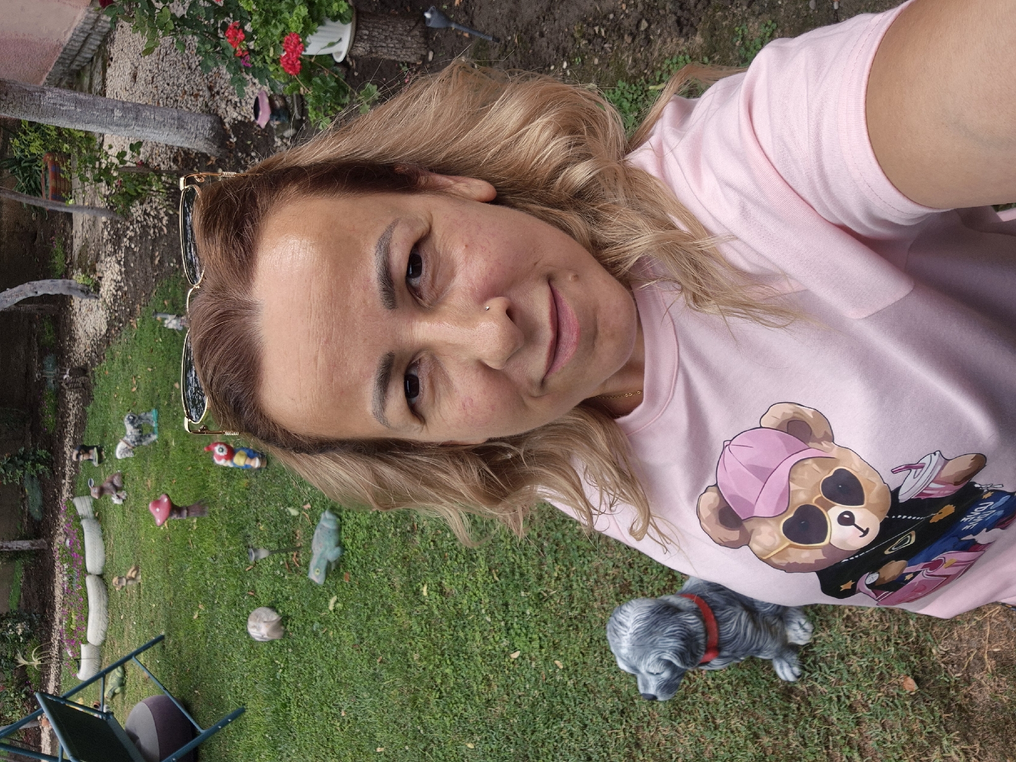 A medium shot shows a fair-skinned woman with shoulder-length blonde hair, wearing a light pink t-shirt with a graphic of a teddy bear wearing sunglasses and a pink baseball cap, and a nose ring. She is standing in a grassy backyard in front of a stone statue of a dog. Various garden ornaments, including a mushroom, frogs, and other animal figures, are scattered across the lawn. In the background, a garden swing set and potted plants are visible. The lighting suggests it is daytime.