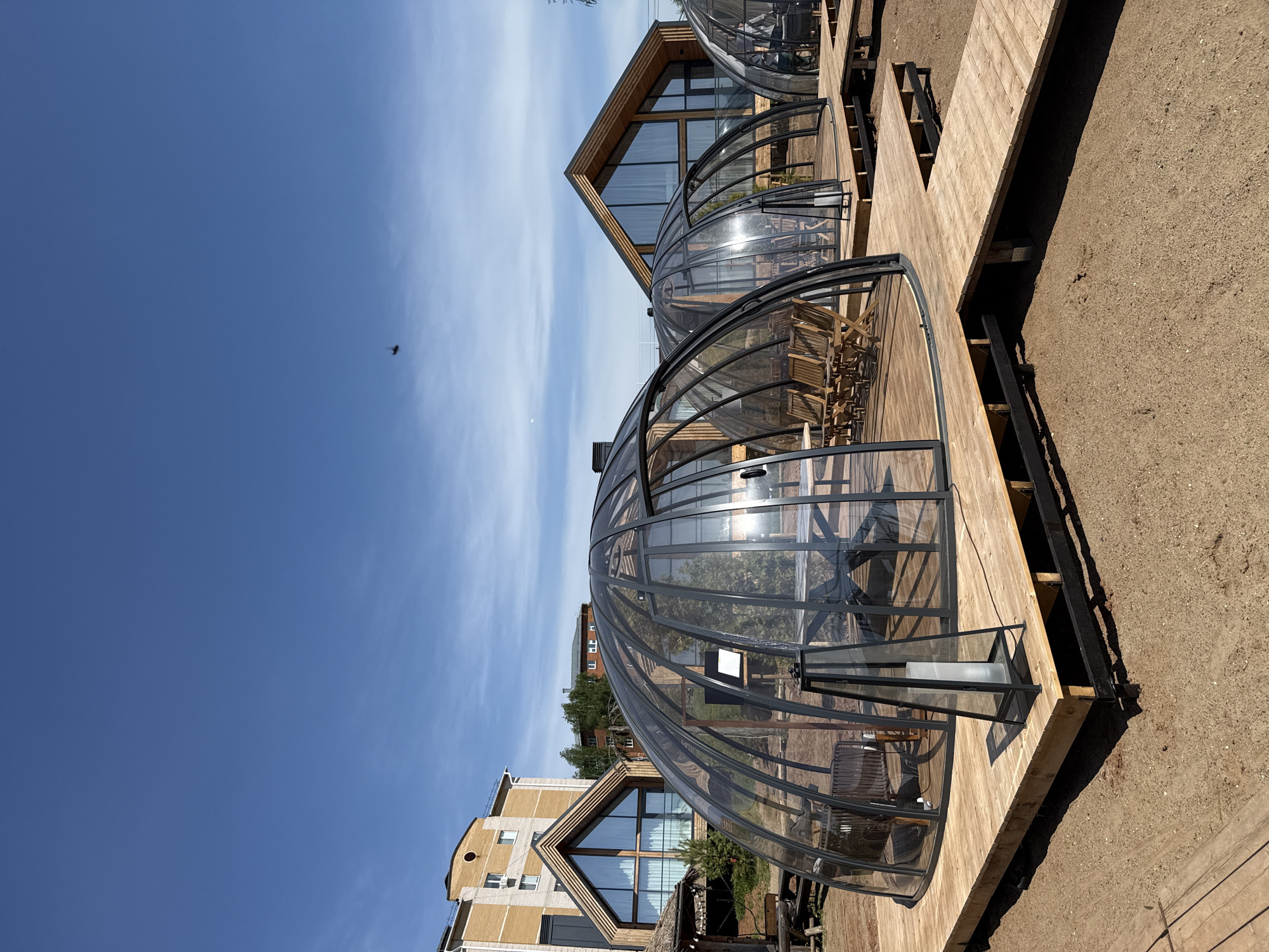 The image shows a ground-level outdoor scene in Votkinsk, Russia, featuring multiple transparent dome-shaped structures situated on a wooden deck. These domes appear to be restaurant or dining spaces, each containing tables and chairs. Adjacent to the domes are buildings constructed with a modern architectural style, incorporating glass and wood elements. The ground surrounding the deck is gravel or dirt. A bird is visible in the clear, blue sky above.