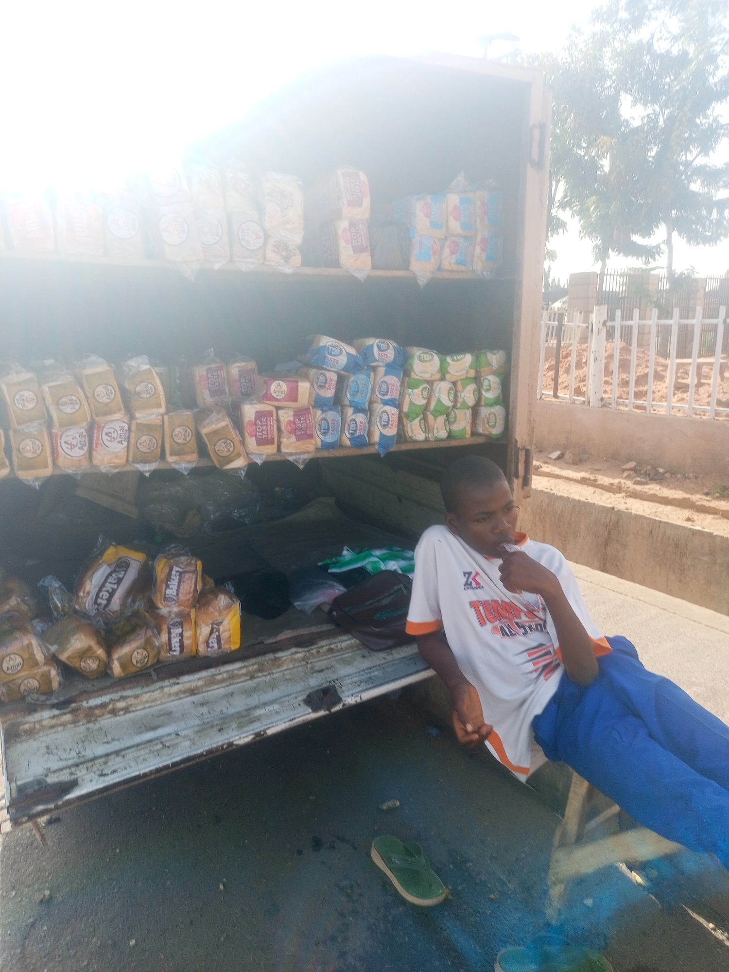 An outdoor photograph shows a bread salesman, a young dark-skinned man, seated in the back of a delivery vehicle on the street in Gombe, Nigeria. The back of the vehicle is open and shelves of loaves of wrapped bread are visible. The brand “TOP TASTE” and “Amal Bakery” are visible on many loaves of bread. The man wears a white t-shirt with orange trim and blue pants. He is seated with his legs stretched out. The road and curb are visible, along with a fence, some buildings, and a tree in the background. The sky is bright.