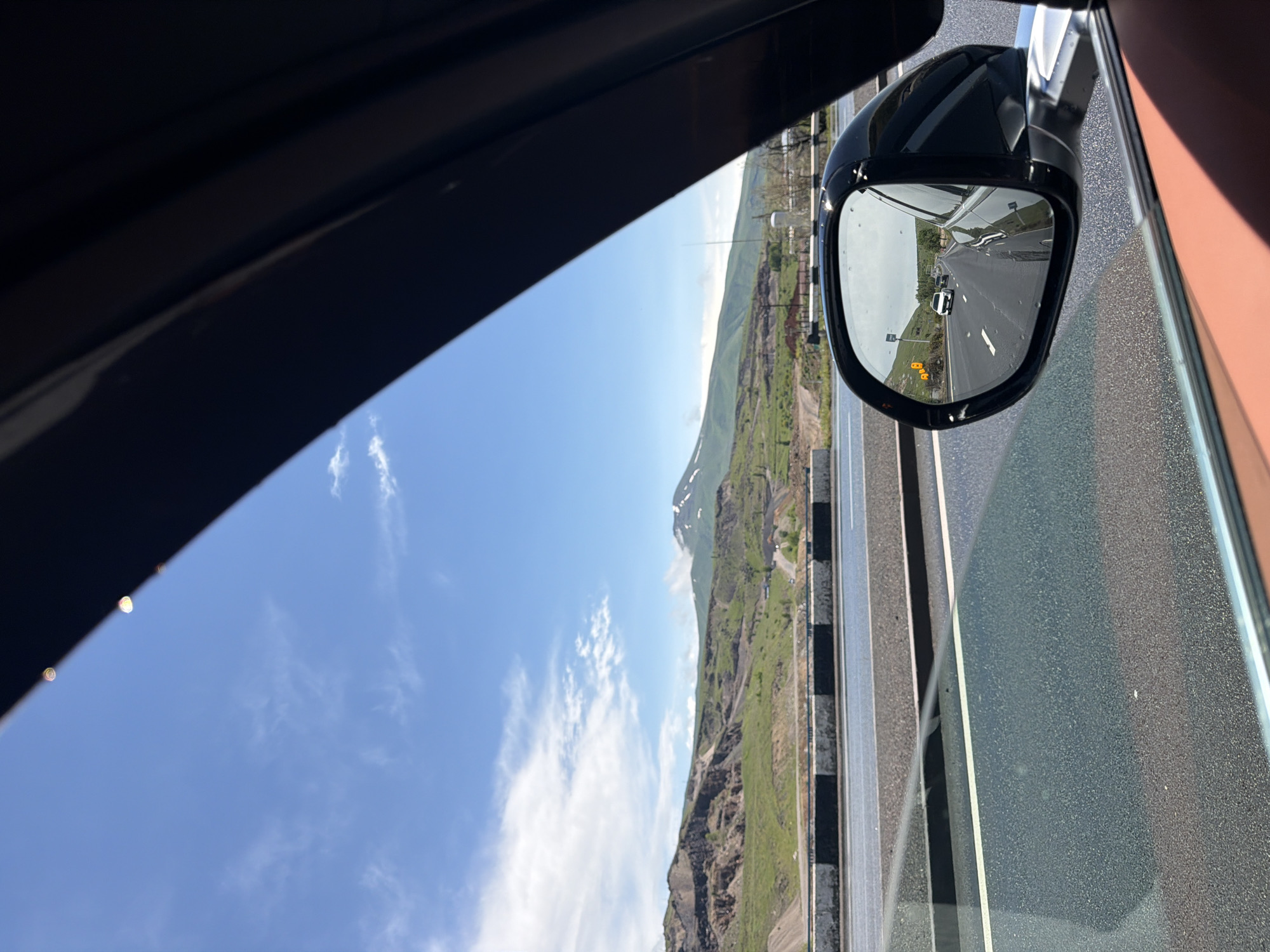 The image shows a view from inside a car. The viewer is presumably on a highway, with a landscape of green hills and mountains visible on the left through the car window. The side mirror of the car reflects a view of the road behind the car, with vehicles traveling on it. The sky is blue with some clouds.