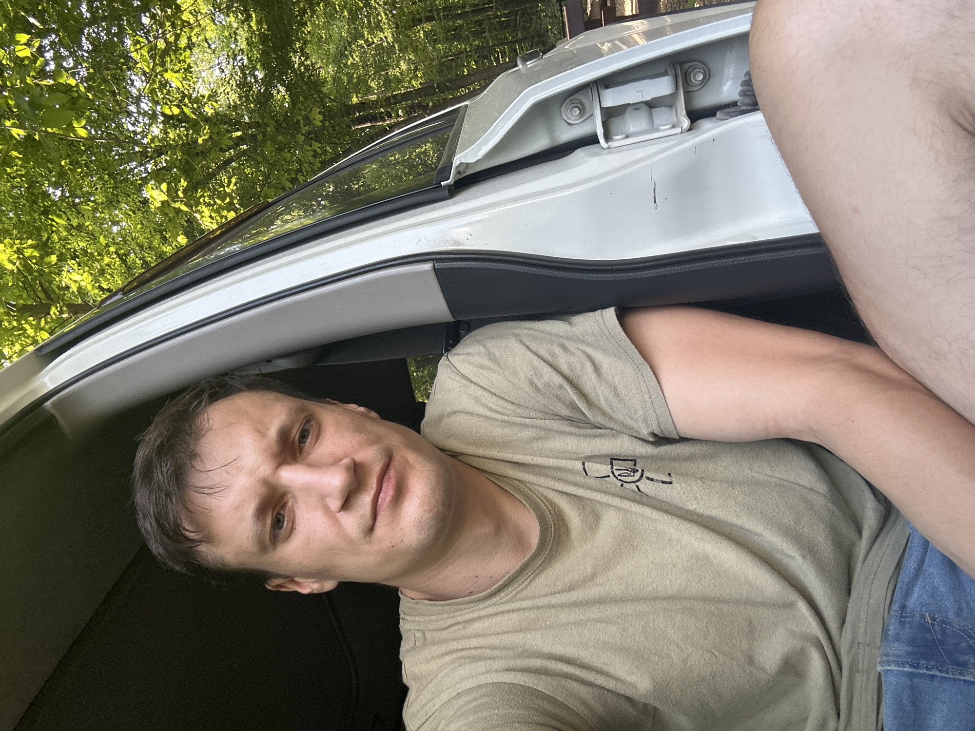 A man with short brown hair and light skin is sitting in the open doorway of a car. He is wearing a light green t-shirt and blue denim shorts. The car door is white and the interior is dark grey. The background is a forest with lush green trees and sunlight filtering through the leaves. The setting appears to be rural, possibly in or near Shatava, Ukraine. The man is looking directly at the camera with a neutral expression.