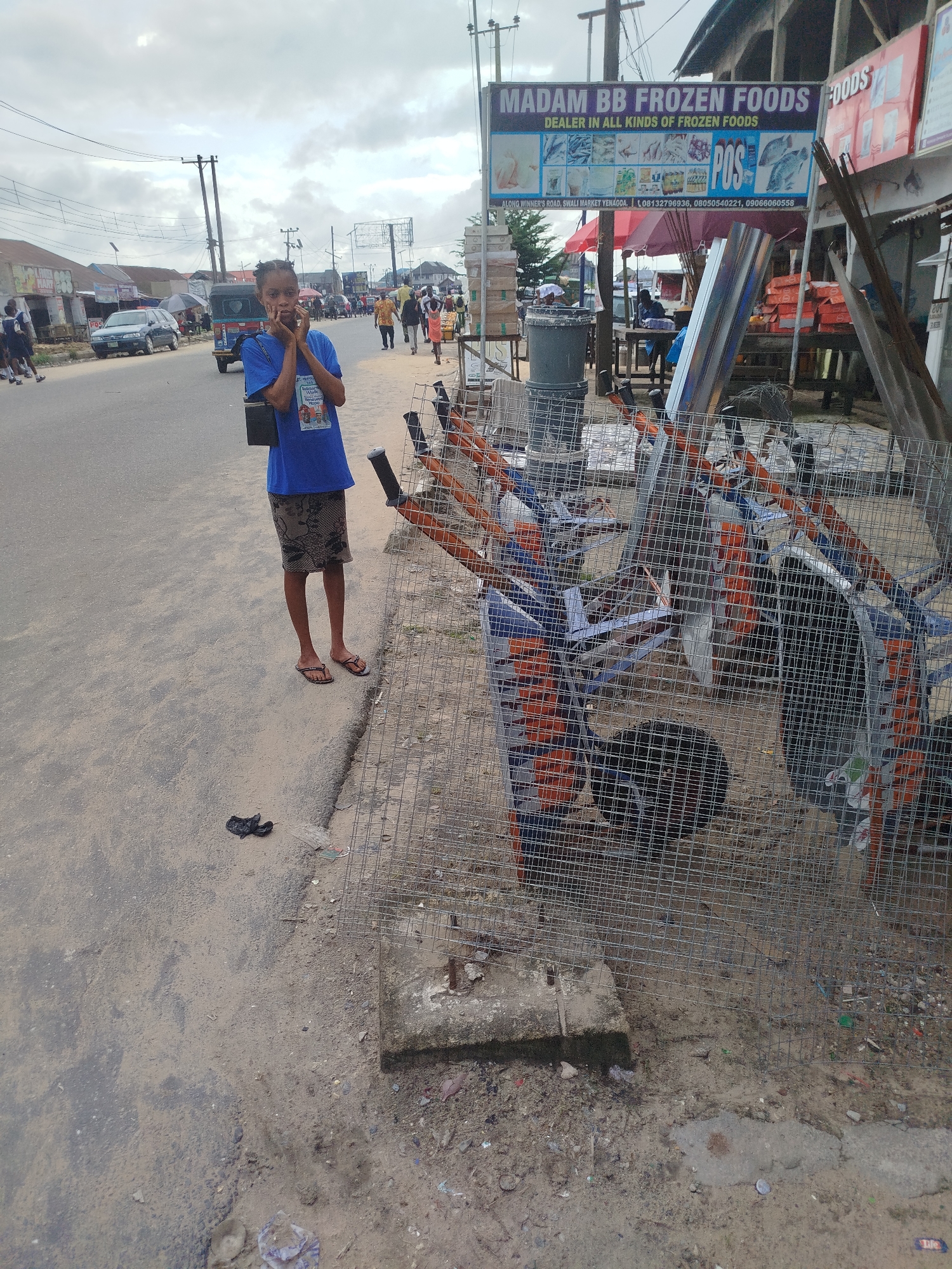 A young girl stands on a sandy road, her hands touching her cheeks as if in thought. She wears a blue t-shirt with an image on it and a patterned skirt, along with flip-flops. In the background, a busy street scene unfolds in Yenegoa, Nigeria. Cars, including a dark SUV and a tricycle, are parked or driving on the road. Various shops and stalls line the street, with a prominent sign for "MADAM BB FROZEN FOODS" indicating a food vendor. The sky is overcast, suggesting a cloudy day. In front of the frozen foods stall, metal cages or enclosures are set up, possibly for displaying or transporting goods. The ground is sandy and somewhat littered, typical of a street market environment. The overall impression is a snapshot of everyday life in a bustling Nigerian town.