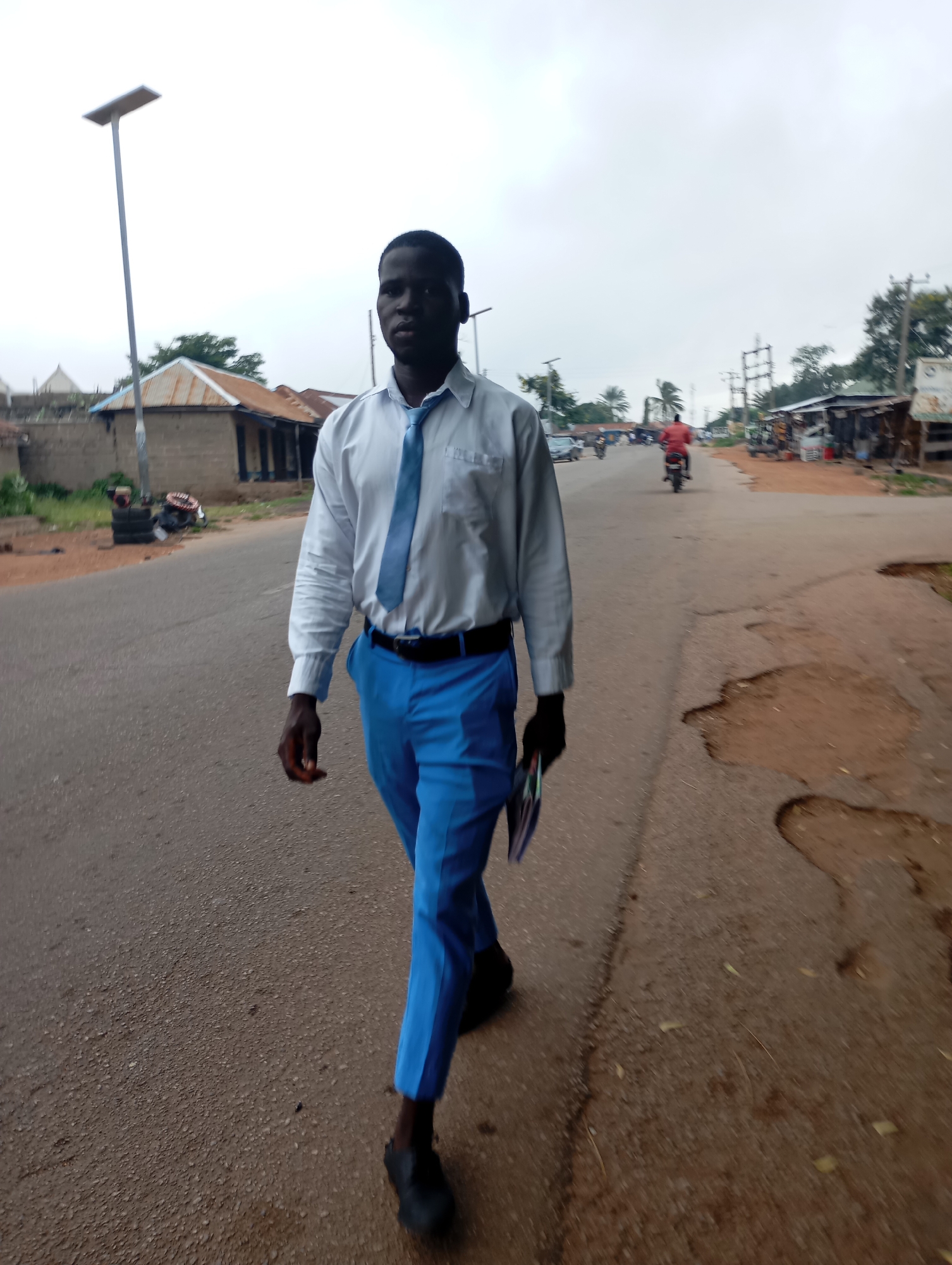 A young African male in a white dress shirt and blue trousers with a blue tie walks along a paved road in Shendam, Nigeria. He is carrying a pamphlet or booklet in his right hand. The road has several potholes and is lined with buildings on the left and shops and vegetation on the right. A solar-powered street lamp stands on the left side of the road. In the background, several people are visible on motorbikes, and cars are present on the road. The sky is overcast.