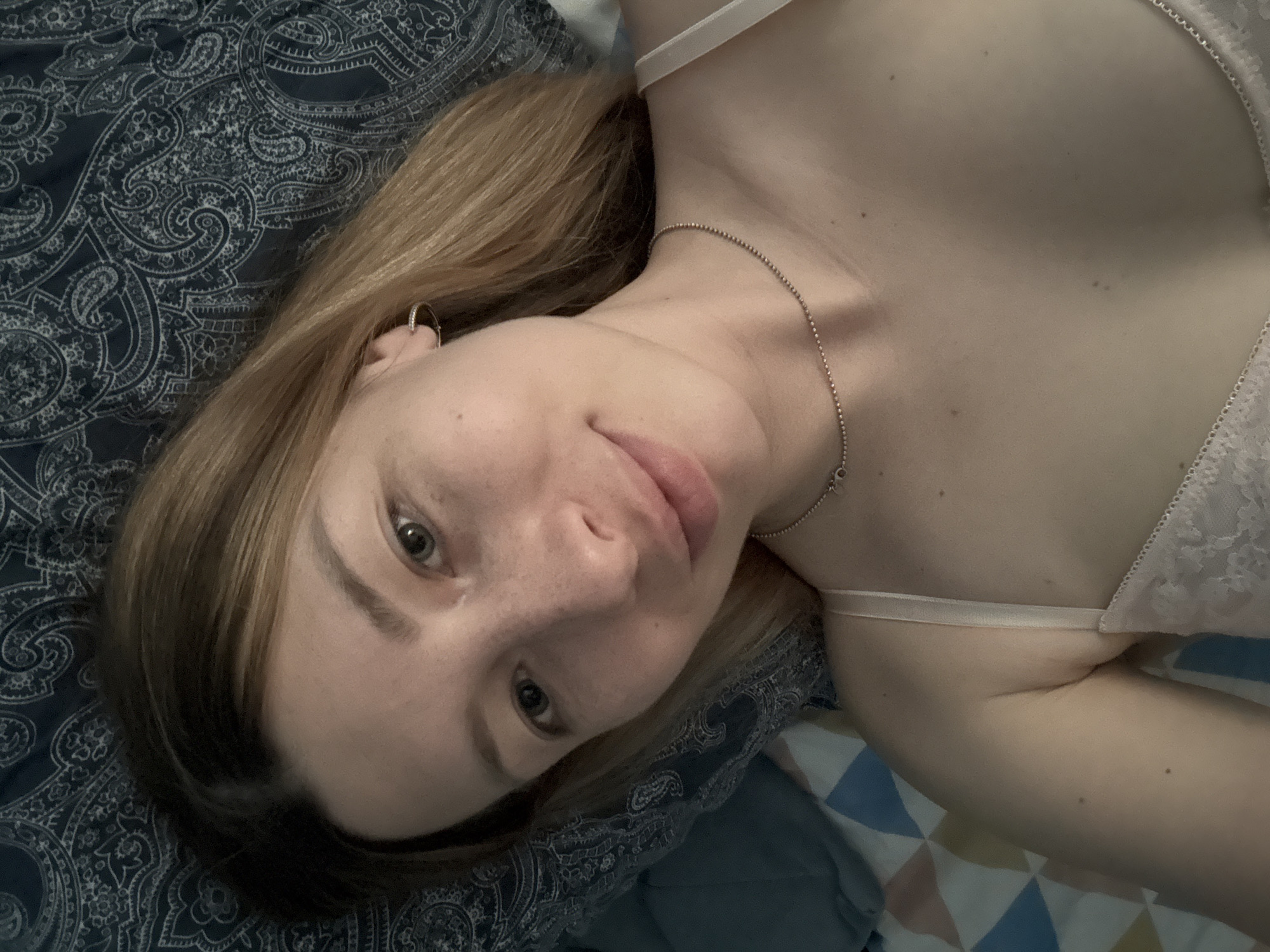 A woman is lying on her back on a paisley-patterned bedspread. She is wearing a light-colored bra and a silver necklace. She is looking at the camera. Her hair is blonde and shoulder-length. There is no indication of a city or location.