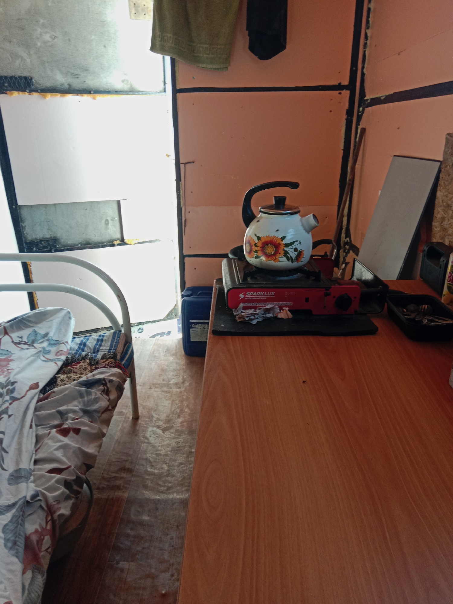 In a simple room, likely a makeshift living space in Akzhar, Kazakhstan, a white kettle with a sunflower design sits atop a portable red and black gas stove. The stove, labeled "SparkLux," is positioned on a wooden table. To the left of the stove, a blue plastic container with a label is visible. Further to the left, a white metal bed frame is covered with a patterned comforter and striped sheets. The room has orange walls with black trim, and the floor appears to be made of wood-look linoleum. A doorway leads to a brighter area, possibly outdoors. A thin wooden stick leans against the wall behind the stove. A dark object, perhaps a towel or cloth, hangs from the ceiling on the upper left. The overall impression is of a basic, utilitarian living area.