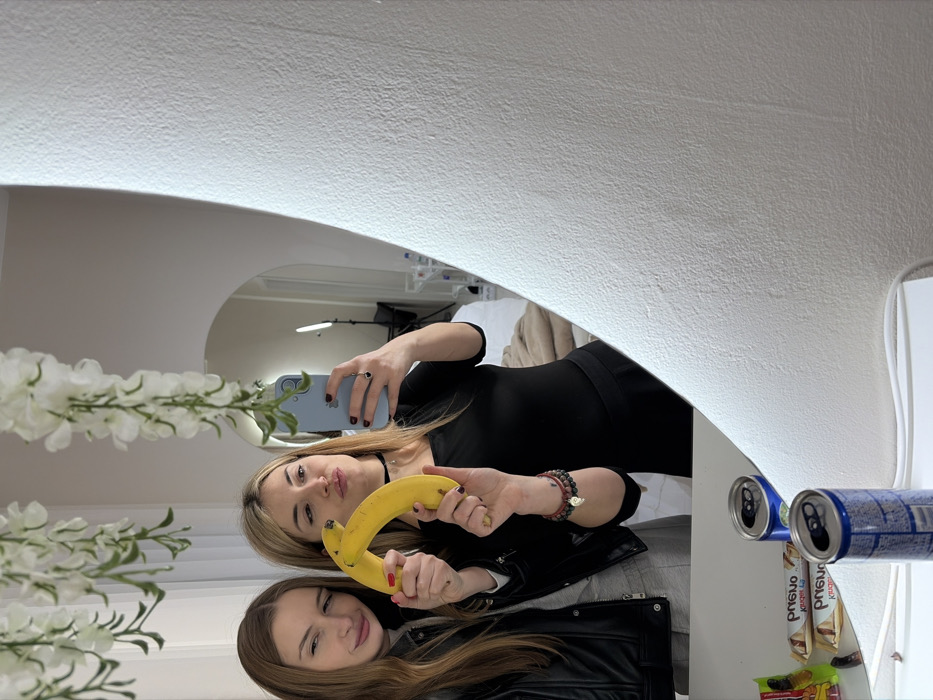 An indoor setting in Budva, Montenegro, captured as a mirror selfie. Two adult females are reflected in a large, irregularly shaped mirror. The female on the left, with long brown hair, wears a black leather jacket and winks, while her right hand holds one end of a yellow banana. The female on the right, with blond hair, wears a black top, has red fingernails, and holds a light blue smartphone in her left hand, operating the camera. Her right hand holds the other end of the banana, and she is puckering her lips. White artificial wisteria-like garlands hang from the ceiling above the mirror. The reflection in the mirror reveals a minimalist room with an arched doorway, two vertical LED light strips, and a light-colored bed or couch with a beige blanket. Below the mirror, on a white surface, are three aluminum beverage cans (two open, one partially visible) and several packaged snacks, including white "Kruger Prijatelj" branded chocolate bars. The wall surrounding the mirror is white and textured.