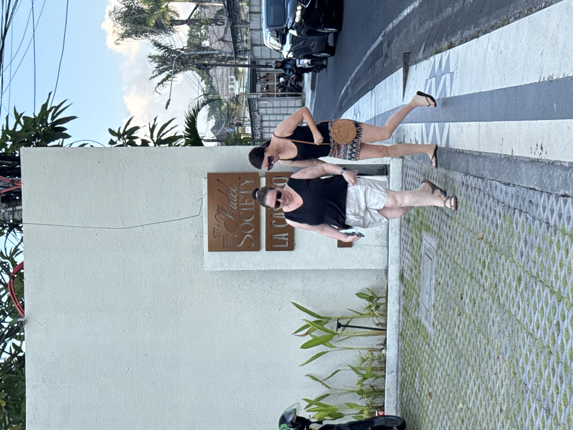 This image depicts an outdoor street scene in Kuta, Indonesia, featuring two women walking on a sidewalk. The time of day appears to be bright daylight, with a clear blue sky partially covered by white clouds.
The woman in the foreground is wearing a black t-shirt, light-colored shorts (possibly white or beige), sunglasses, and flip-flops. She holds a phone or small object in her left hand. The second woman, walking slightly behind and to her right, is dressed in a black tank top, patterned shorts (with a geometric design), sunglasses, and black flip-flops. She carries a round, woven straw bag with a long strap across her body. Both women appear relaxed and are casually dressed, suggesting a leisurely stroll.
They are walking alongside a light-colored, possibly white, building. On the building's facade, two rectangular plaques are mounted vertically. The upper plaque clearly displays "The Palace Society", while the lower one shows "LA CANTINA". In front of the building and along the sidewalk, green plants are growing, likely in planters.
To the right of the sidewalk is a street with a pedestrian crossing marked by alternating gray and white stripes. A black car is visible on the street further in the background, and several motorcycles or scooters are parked along the roadside. Overhead, numerous utility wires crisscross against the sky. Tropical foliage, including palm trees, is scattered throughout the background, indicative of the Indonesian setting. The sidewalk is made of patterned concrete pavers with grass growing in the gaps. A green and black scooter helmet is partially visible in the lower-left corner. The overall scene conveys a typical sunny day in a lively urban environment.