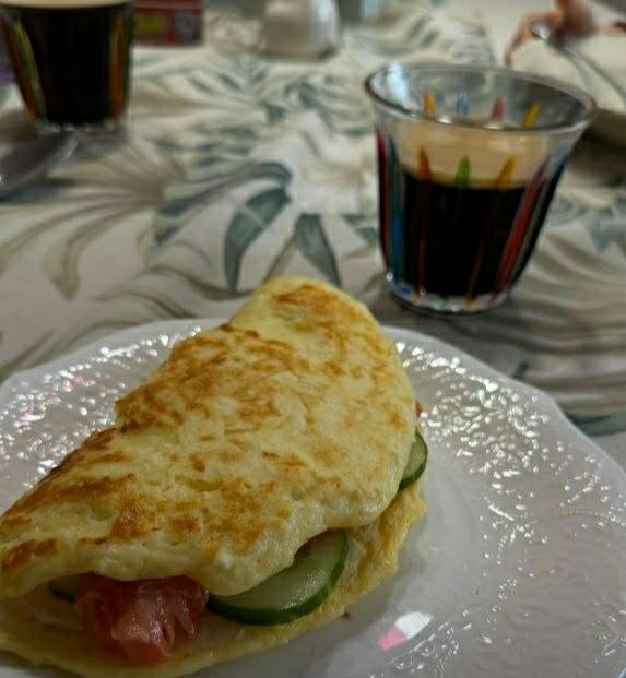 A close-up shot shows an omelet filled with salmon and cucumber slices on a white plate, with a glass of coffee and another container in the background. The environment suggests an indoor breakfast setting. The location is not identifiable from the image.