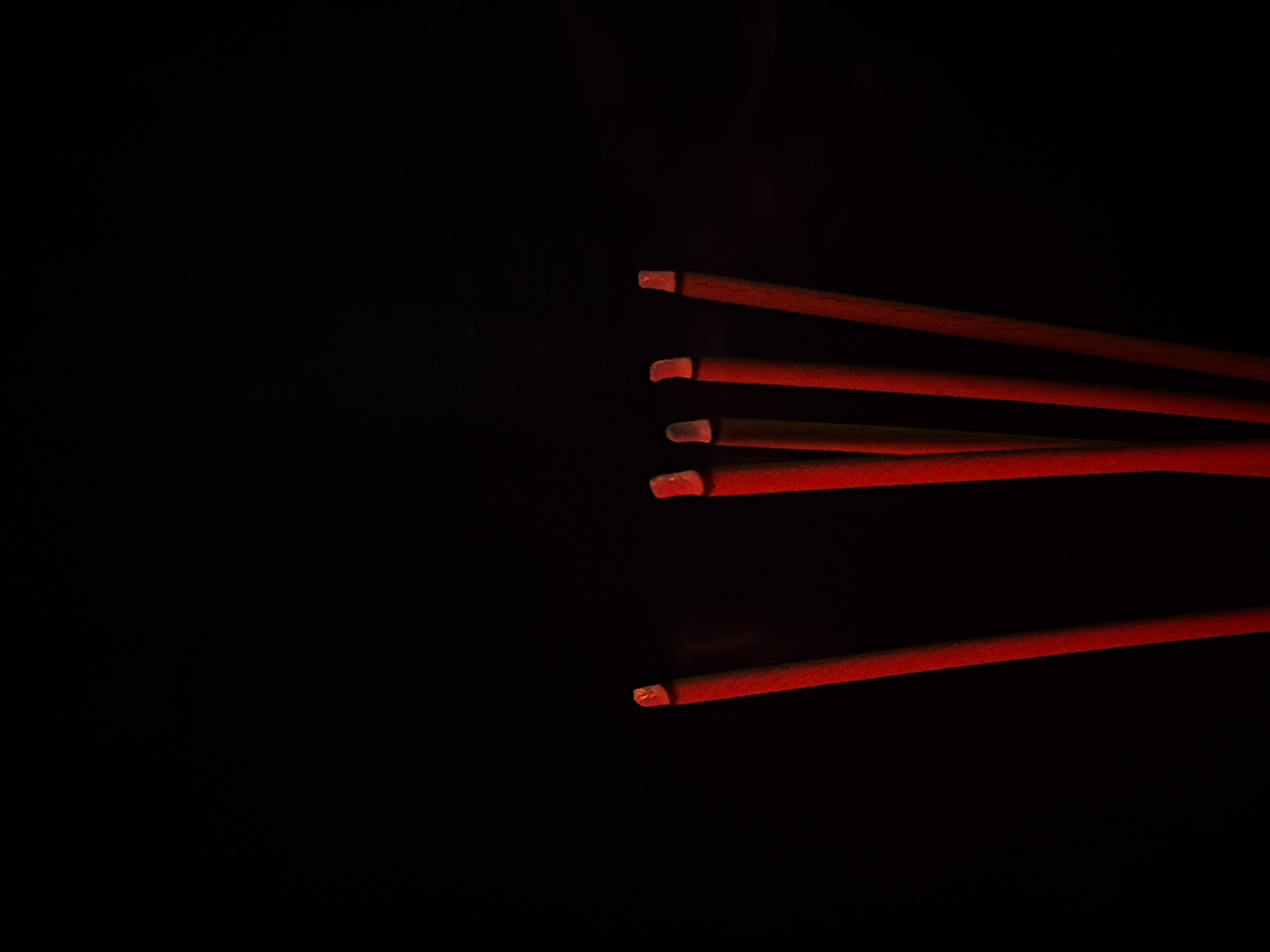 A close-up, low-angle shot captures a cluster of five red incense sticks against a solid black background. The tips of the incense sticks are glowing red, indicating they are burning. A faint wisp of smoke is visible rising from the leftmost incense stick. The composition is centered on the incense sticks, with the majority of the frame dedicated to the dark background.