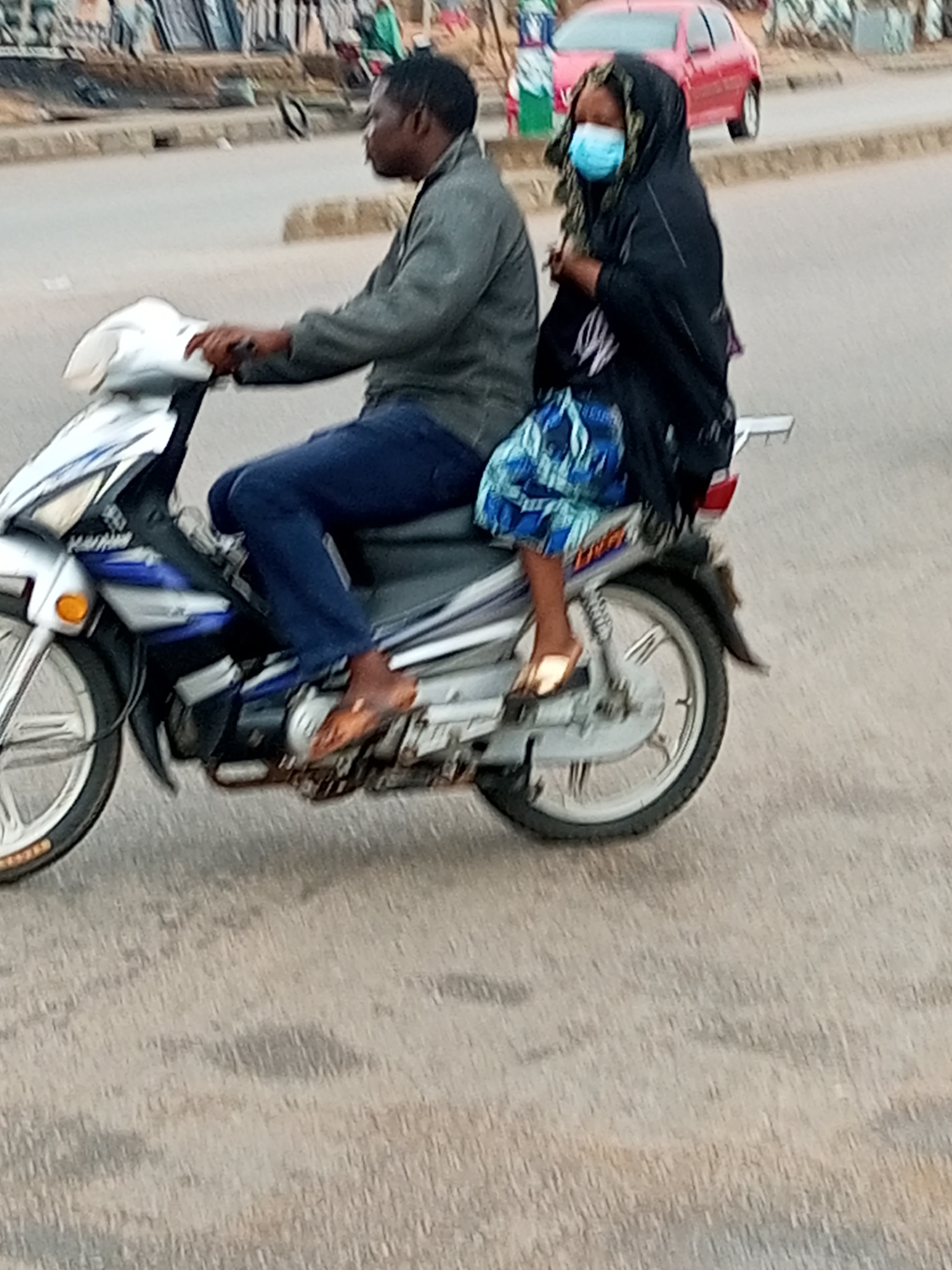 In Gombe, Nigeria, a man and a woman ride on a motorcycle on a paved road. The man, wearing a gray jacket and dark blue pants, is driving. The woman, wearing a dark hijab and a blue and white patterned skirt, is the passenger. She is also wearing a light blue face mask. The motorcycle is silver and blue. The background shows a blurry urban environment with some buildings and a red car. The road surface is asphalt.