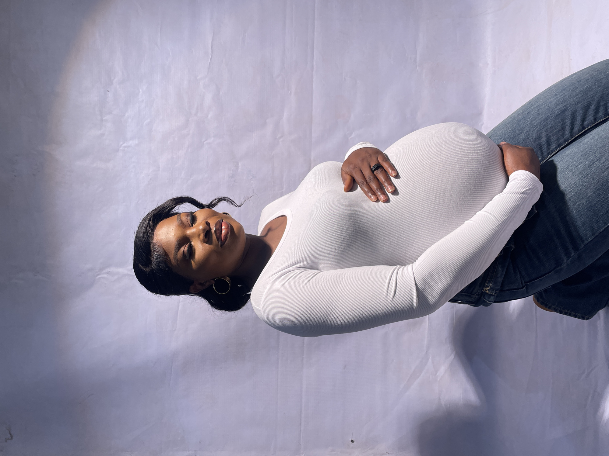 A pregnant, dark-skinned woman wearing a white, long-sleeved shirt and blue jeans lies on her back against a white backdrop. Her hands are placed on her pregnant belly. She wears a black ring on her right hand and gold hoop earrings. Her hair is styled back from her face. The image provides no information to identify the location as Jos, Nigeria, or any other city.