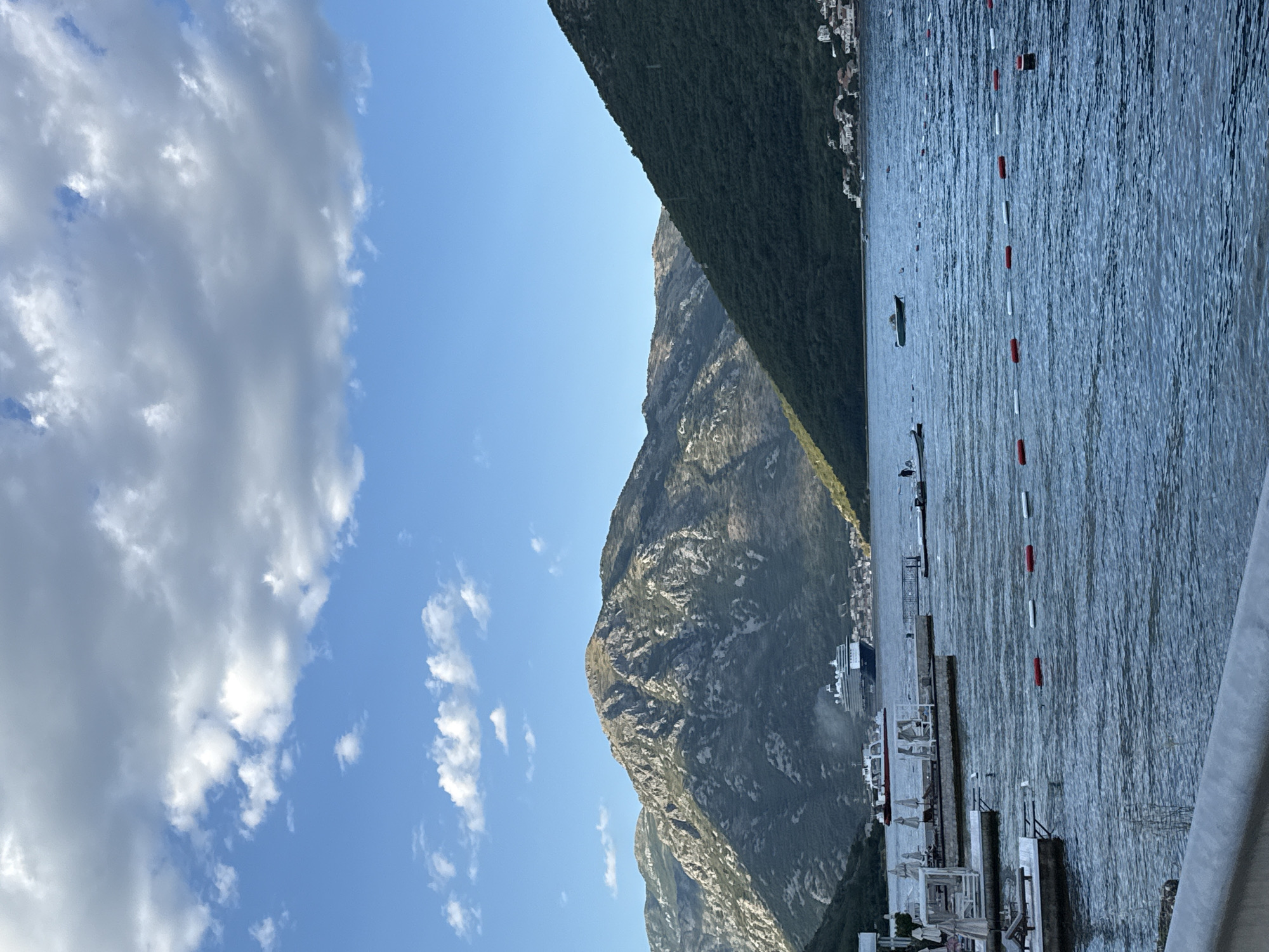 This is a vertical, eye-level, full shot of a body of water with mountains in the background. The water is dark blue and choppy, with a few buoys and boats floating on it. In the background, there are large, rugged mountains covered in greenery. A cruise ship and a ferry are visible on the water, and some buildings are nestled into the mountainside. The sky is blue with scattered white clouds. A pier with lounge chairs and umbrellas is in the foreground on the left. The scene appears to be in a bay or fjord, with mountainous terrain and water. The location is Jošice, Montenegro.