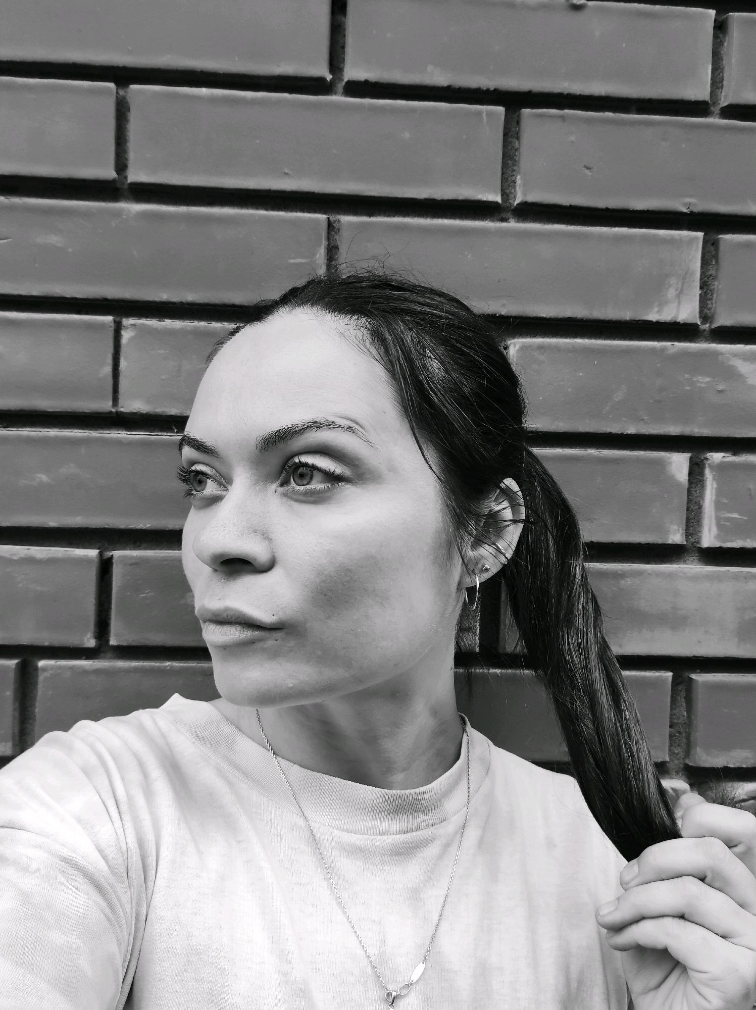 A black and white, medium shot photograph captures a woman in profile, facing left, against a brick wall. She has dark hair pulled back into a ponytail, which she is holding with her right hand. She is wearing a light-colored t-shirt and a delicate necklace. Her facial expression is neutral, with her gaze directed off-camera. The brick wall behind her is constructed of rectangular red bricks, laid horizontally. The image appears to have been taken outdoors in an urban setting. No specific landmarks or distinguishing features of the city of Фоки, Russia, are visible in the photograph.