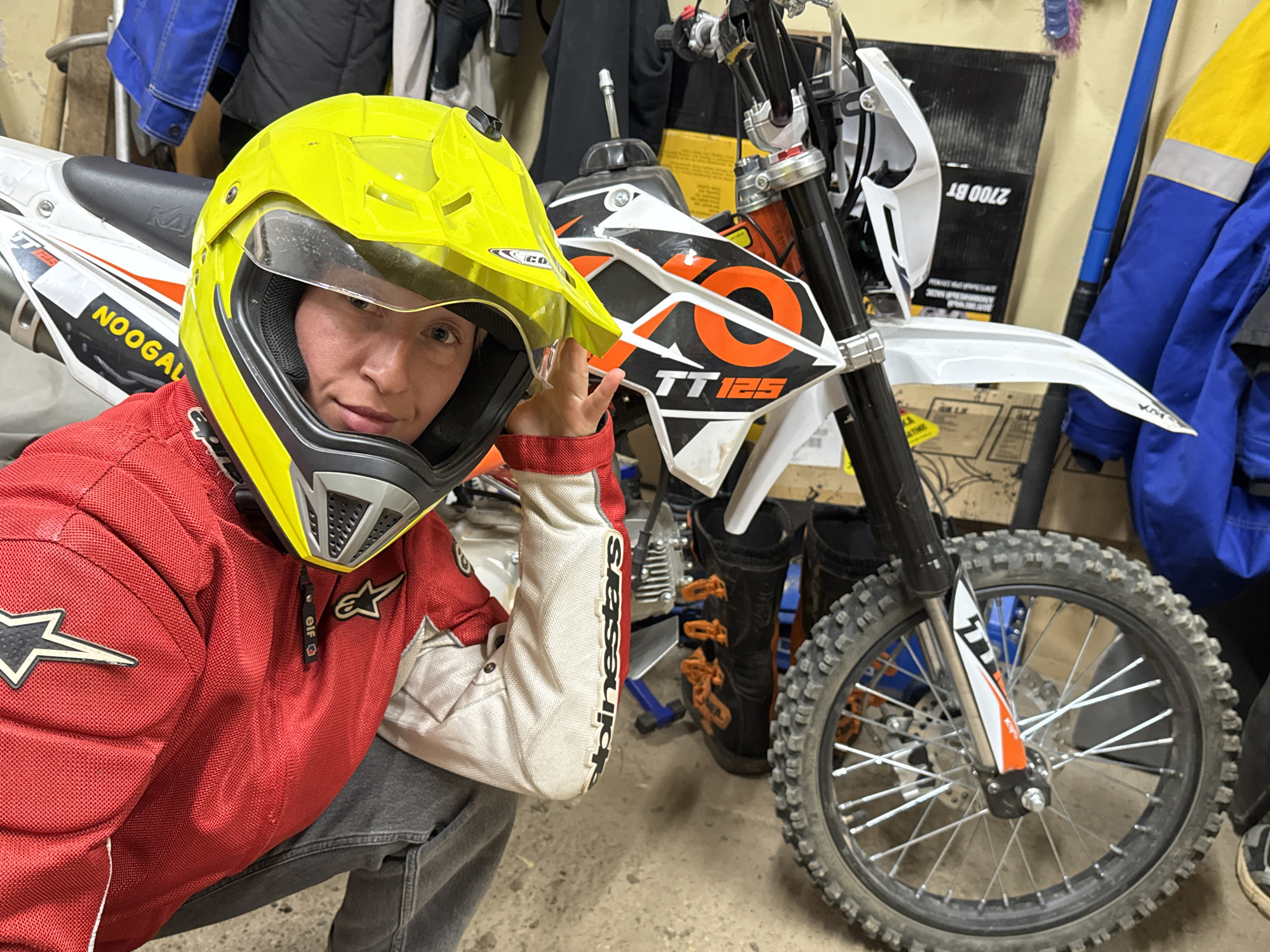 A person wearing a yellow helmet, red and white racing jacket, and gray pants is crouched next to a white and black dirt bike with an orange "O" design and "TT125" printed on the side. The dirt bike has a white front fender, a black front tire, and orange-accented boots behind the tire. The person is holding their hand near their face. The background includes a wall with hanging jackets, a cardboard box, and other miscellaneous items. The scene appears to be in a garage or storage area with a concrete floor. The location in the city of Chaikovsky, Russia, is not identifiable from the image alone.