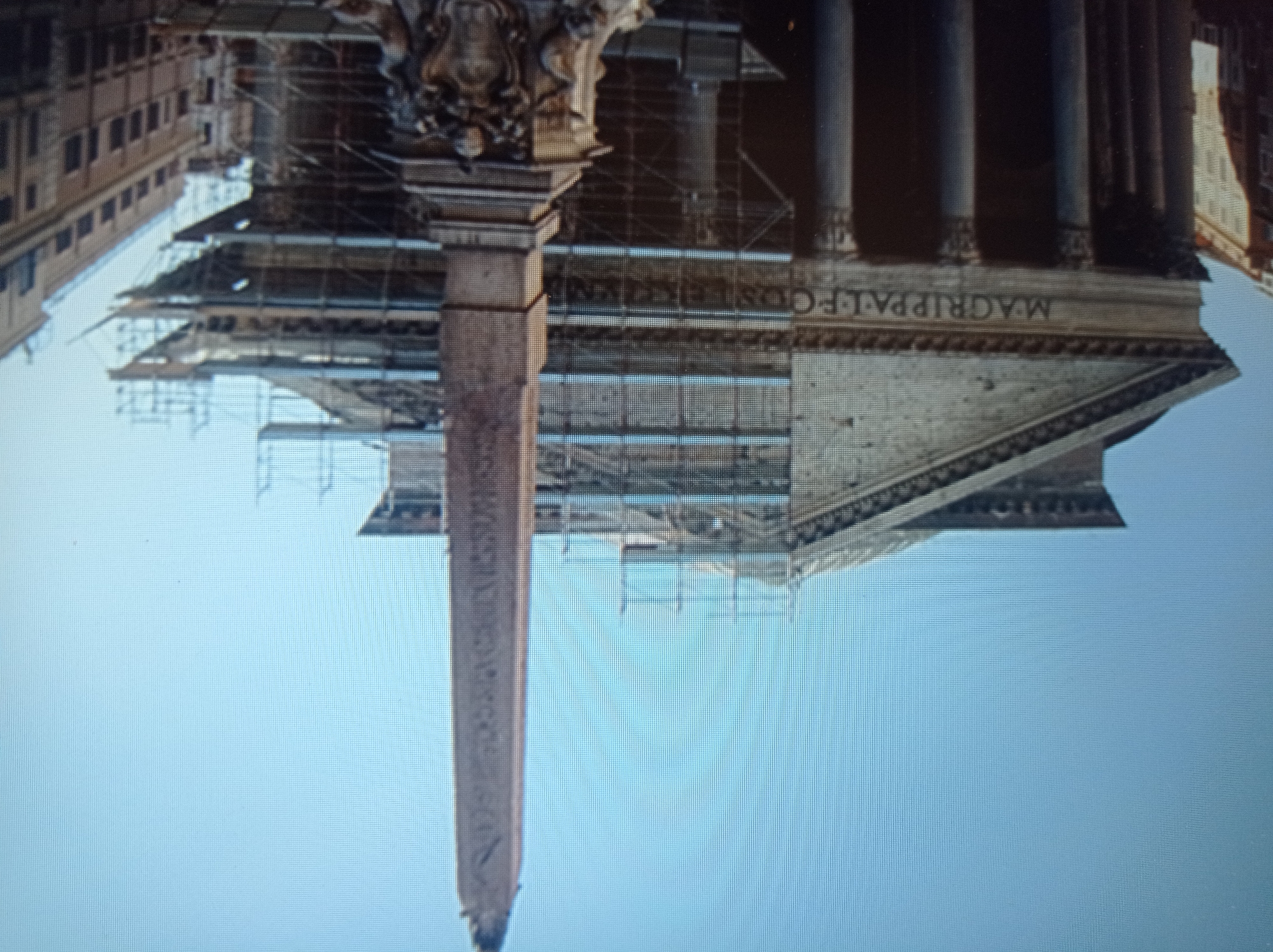 The image depicts an ancient structure, likely a temple or monument, with a large obelisk in the foreground. The building features classical columns and a triangular pediment. Scaffolding is visible around the structure, suggesting ongoing restoration or maintenance work. The sky is clear and blue. The inscription "M. AGRIPPA L. F. COS. TERTIUM FECIT" is visible on the building's facade. To the right of the main structure, a multi-story building with windows is visible. The scene does not contain any elements that suggest a location in Mukachevo, Ukraine.