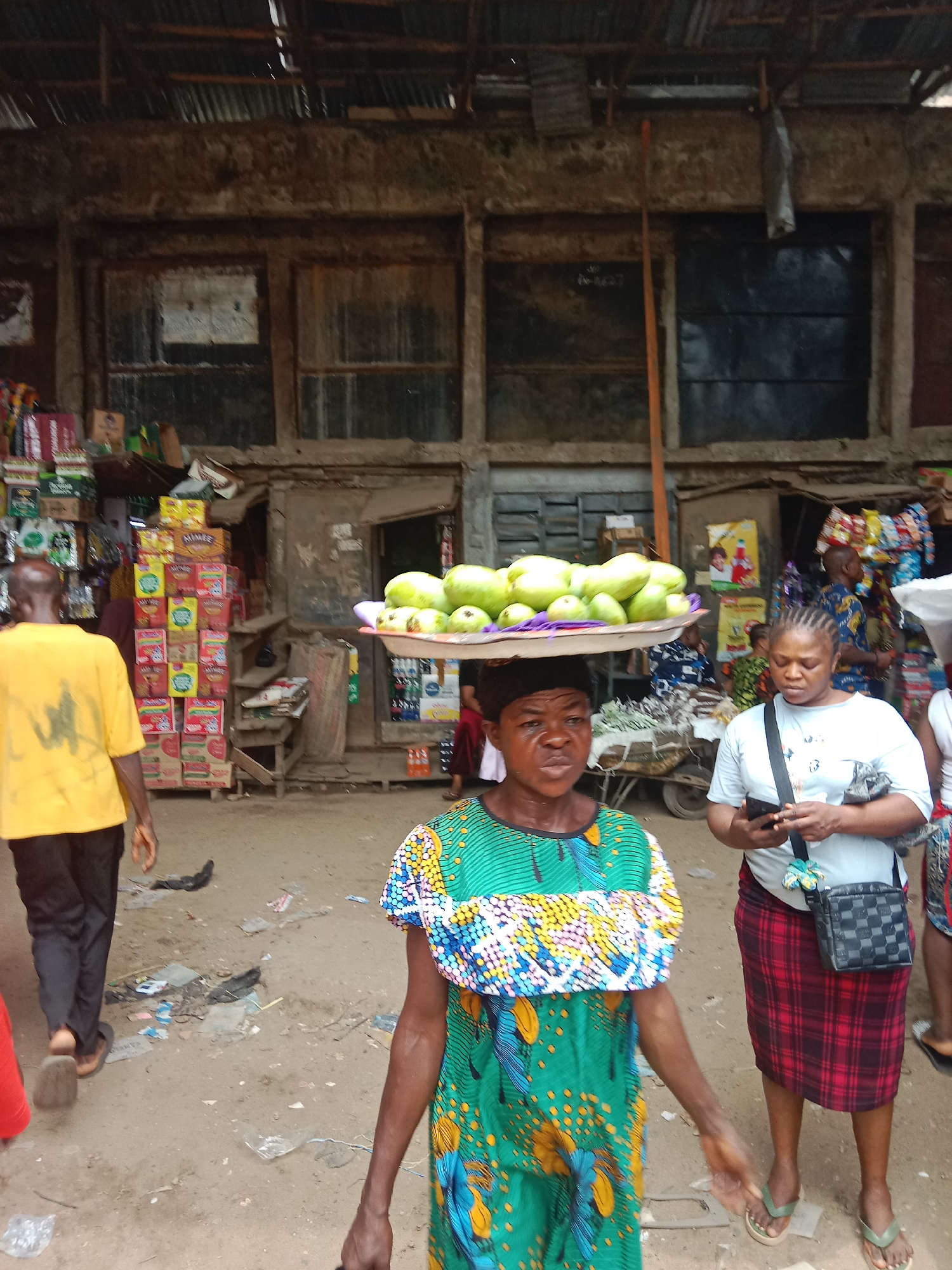 The image depicts a street scene in Onitsha, Nigeria, characterized by a crowded outdoor market environment. A woman in the foreground balances a tray laden with green fruits on her head, her facial expression suggesting concentration. She wears a colorful patterned dress with a green and blue bodice. To her right, another woman checks a mobile phone, dressed in a white t-shirt and a red plaid skirt, with a black checkered shoulder bag.
Behind them, a man in a yellow t-shirt walks away from the camera on the left. Stacks of boxed goods are visible near storefronts. The background shows buildings with weathered facades and corrugated metal roofs. Debris and litter are scattered on the ground, indicative of a busy market area.
