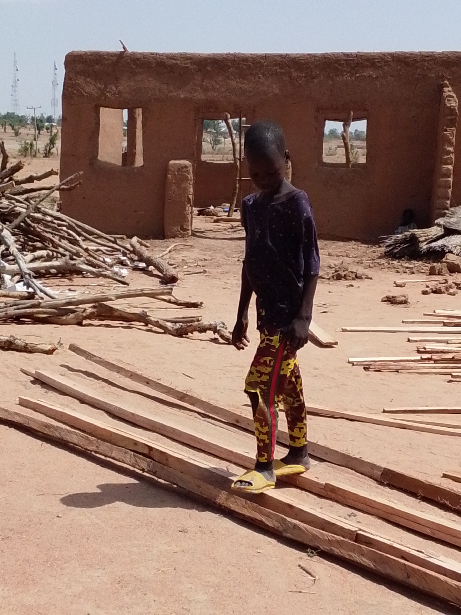 A child walks across planks of wood in a dirt yard. Behind the child is a dilapidated mud brick building with window openings. A pile of branches and some straw-like material are also visible in the yard. Utility poles can be seen in the distance. The location is suggested to be Mulke Barikin Sayejo, Nigeria.