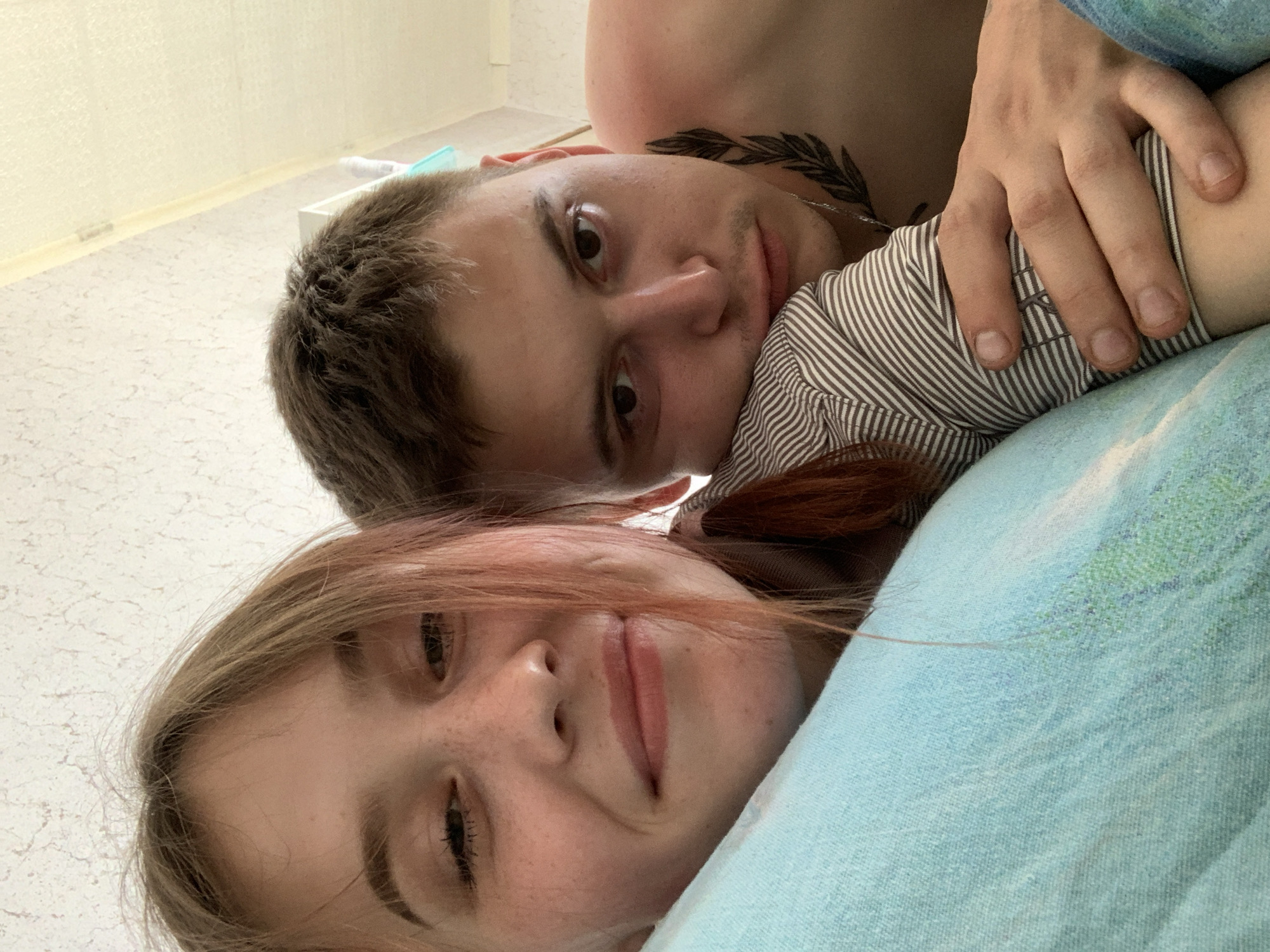 A close-up, tilted shot depicts a young woman and man lying together. The woman, positioned below, faces the camera with a slight smile. She has fair skin with freckles, light brown hair with reddish highlights, and defined eyebrows. Above her, the man looks directly at the camera with a more serious expression. He has dark hair and a tattoo of a leafy branch on his neck. His hand rests on what appears to be the woman's striped-sleeved arm. The background is ambiguous, but it is possible that this photo was taken in an indoor location somewhere in the city of Фоки, Russia, as requested. The setting appears to be intimate, possibly a bed, indicated by the blue-green fabric they are lying on. The image is brightly lit, enhancing the details of their faces and the textures of their clothes and surroundings.