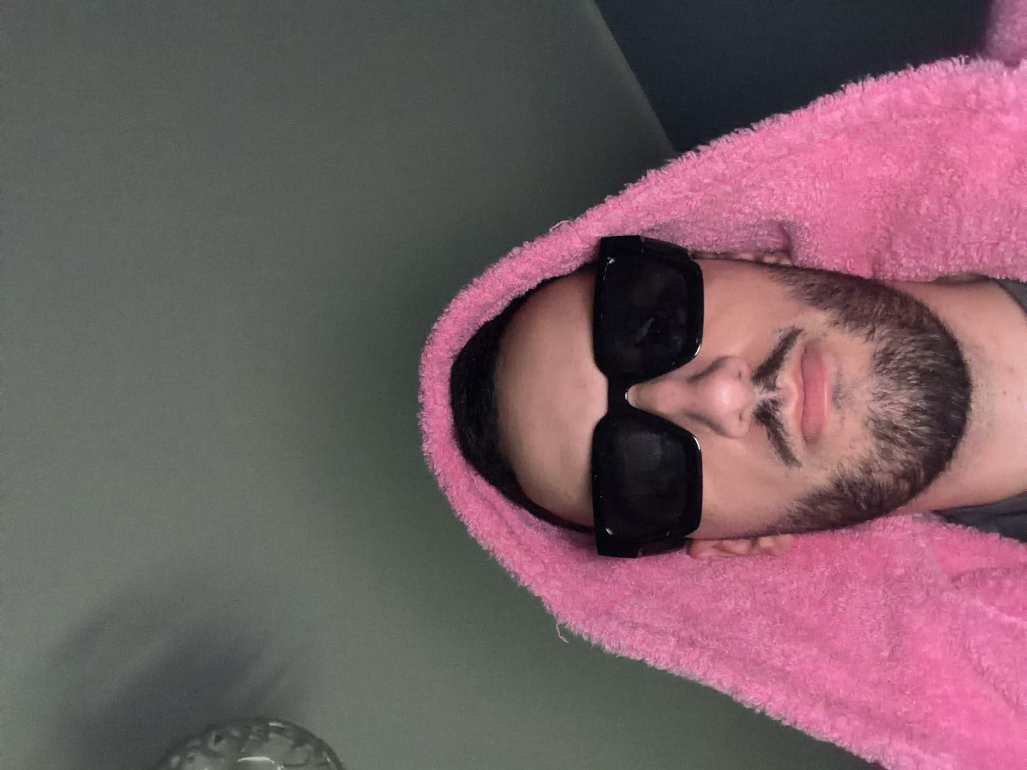A man lies on a light grey surface wearing a pink hooded towel and black sunglasses. The location is unknown and cannot be determined from the image.