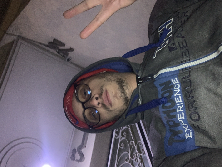 A man in a gray hoodie, red bandana, and round glasses poses indoors with a peace sign. Surveillance cameras are affixed to a door frame in the background. The man has a short beard and mustache. A white door with ornate metal detailing is visible, along with blurred lighting. The hoodie has a blue stripe with "TYPHOON EXPERIENCE" and "FOR RULE BEAKER" printed on it.