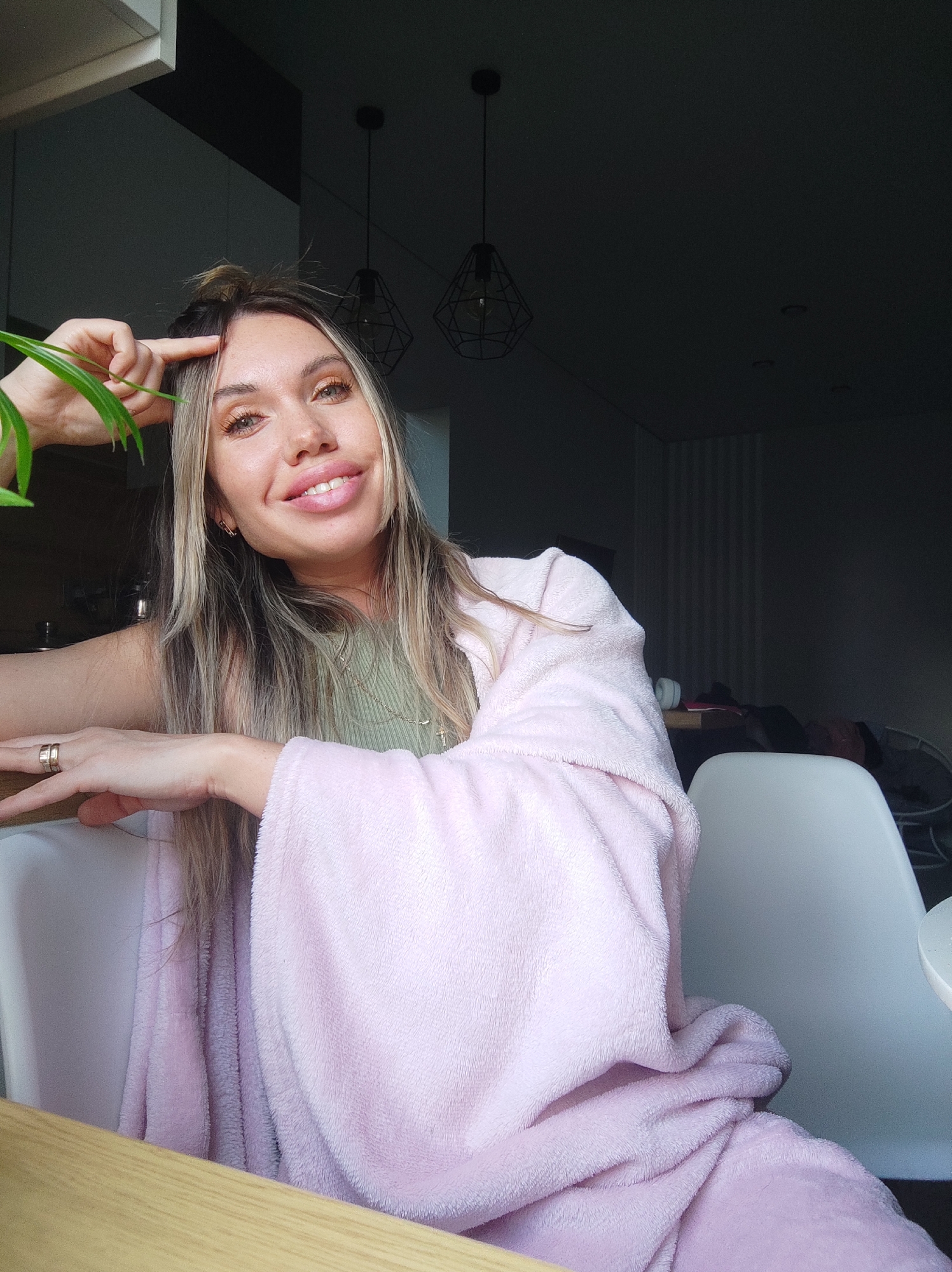 A woman with blonde and light brown hair, styled partially up and flowing over her shoulders, is depicted smiling broadly. She has light-colored eyes with golden eye makeup and full lips. She wears a light green ribbed top and is wrapped in a soft, pink fluffy blanket or robe. Her left hand, adorned with two gold rings, rests on a light wooden surface, possibly a table or counter. Her right hand is raised, with her index finger pointing towards her forehead. A green plant leaf is visible in the upper left corner, close to her face.
The setting appears to be an indoor space, likely a home in Votkinsk, Russia, characterized by dramatic lighting. The foreground features the wooden surface and a white chair back. The background is predominantly dark, creating a strong contrast with the illuminated subject. Two black geometric pendant lights hang from the dark ceiling in the mid-background, and faintly visible recessed lights are also present. Further back on the right, vertical striped wallpaper or a textured wall can be discerned in the dimly lit room, along with parts of other furniture and a second white chair. The activity suggests she is posing casually, possibly for a portrait, conveying a relaxed and happy emotion. There is no visible text in the image.