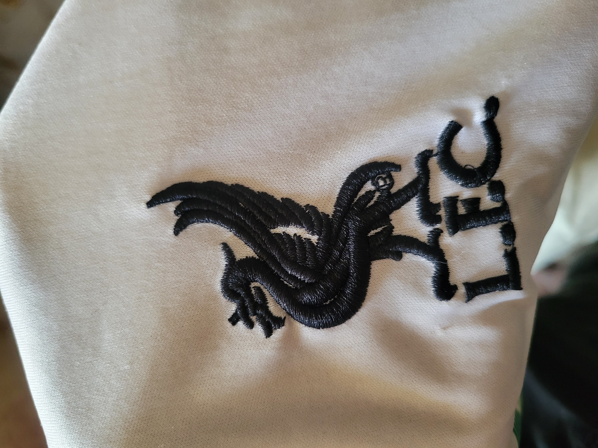 The image depicts a close-up of a white fabric with a black embroidered design. The design includes a Liver Bird, the symbol of the city of Liverpool, England. The abbreviation "LFC" (Liverpool Football Club) is embroidered next to the Liver Bird. The location and time are not ascertainable from this image.