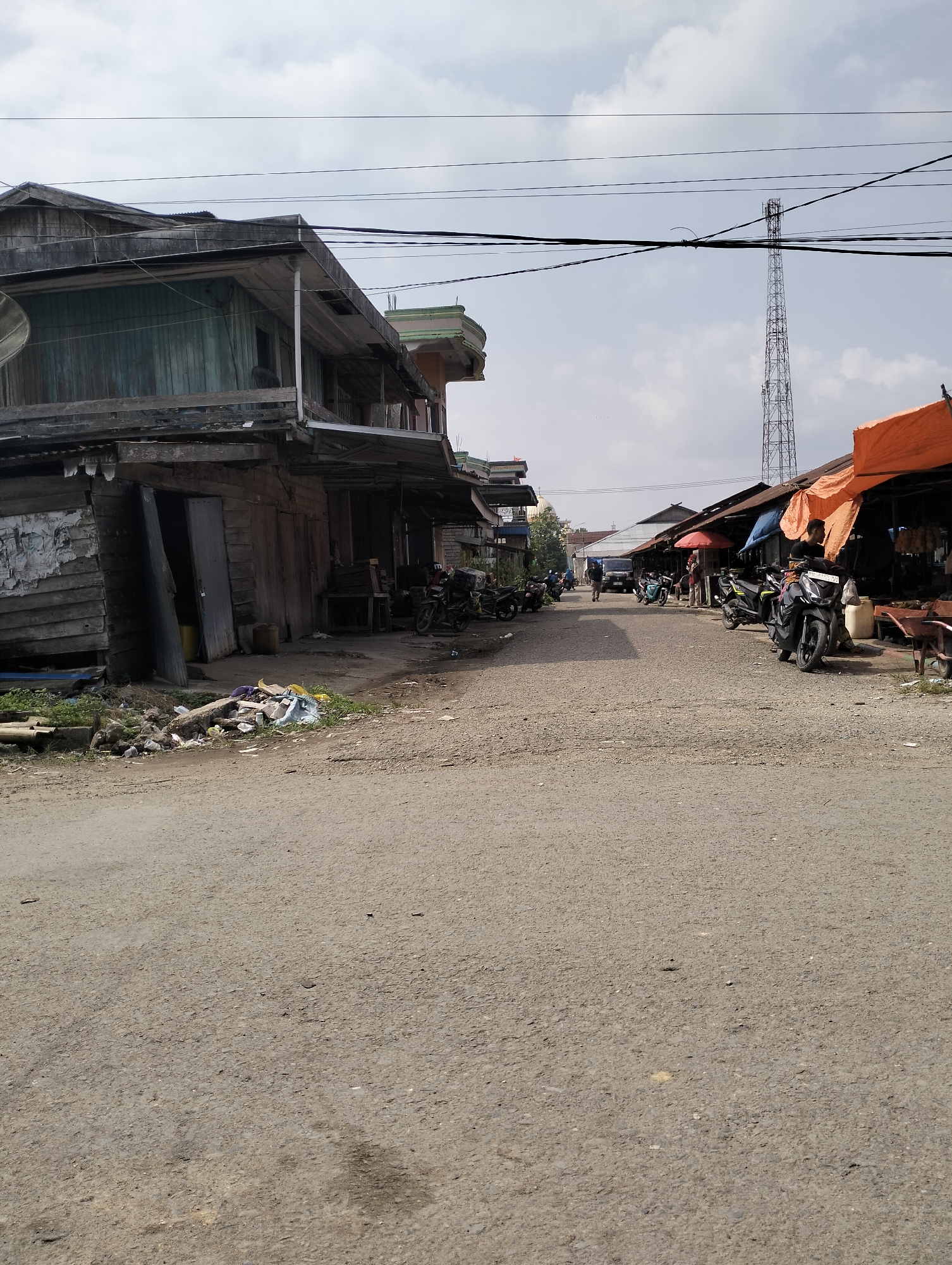 The image depicts a street scene in Serdang Jaya, Indonesia, characterized by a mix of commercial stalls and residential structures. The foreground is an unpaved road surface. On the left side of the street, weathered wooden buildings with open doorways and visible interiors are present. Some of these structures appear to be in disrepair, with peeling paint and exposed wood. Piles of debris and vegetation are visible at the base of these buildings.

Further down the street, commercial stalls with orange and blue awnings are set up, suggesting a marketplace. Several motorcycles are parked along the street and in front of the stalls, indicating active commerce or transportation. People are visible in the distance, some standing near the stalls and one individual riding a motorcycle. A tall communication tower stands prominently in the background, against a cloudy sky. The overall impression is of a functional, albeit somewhat aged, urban or semi-urban environment in Indonesia.